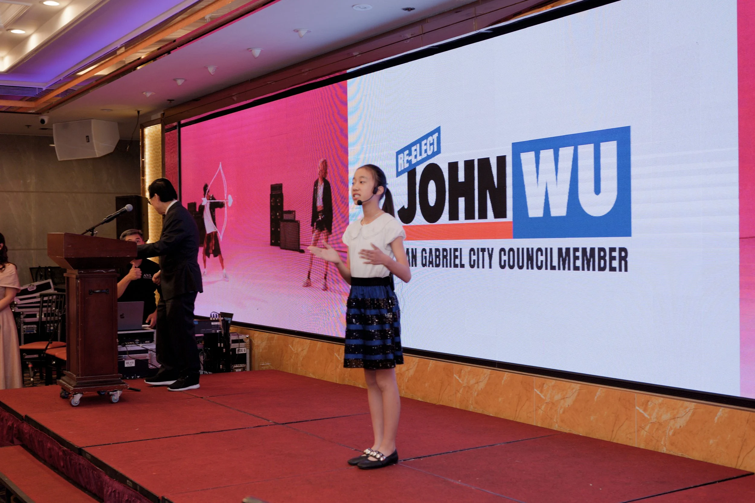 A young girl with a microphone headset standing on a stage in front of a large screen displaying the name John Wu and the title City Council Member, with a man speaking at a podium and others seated nearby.