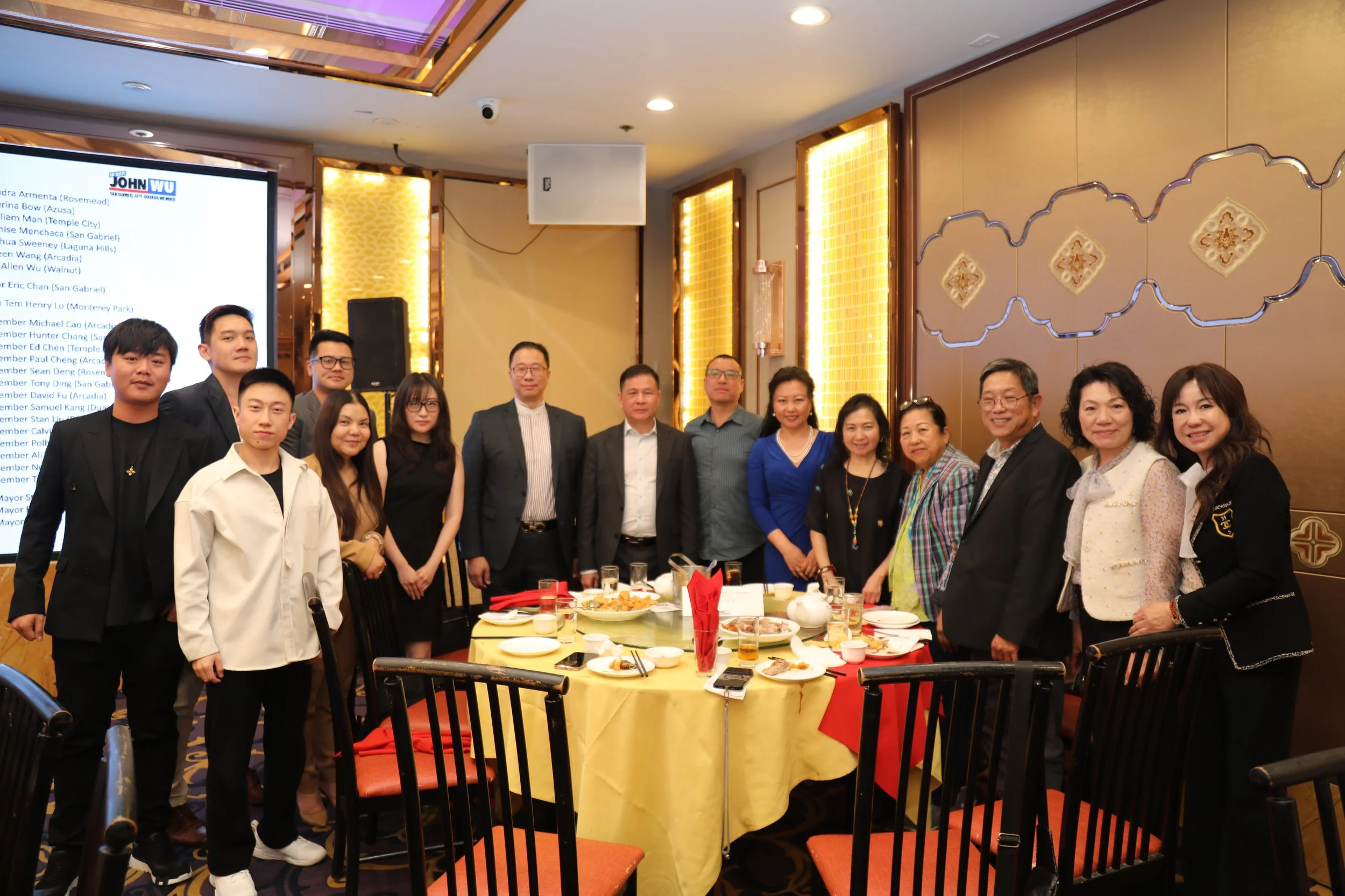 A group of people standing around a table with food at a celebration or dinner event in a decorated banquet hall.