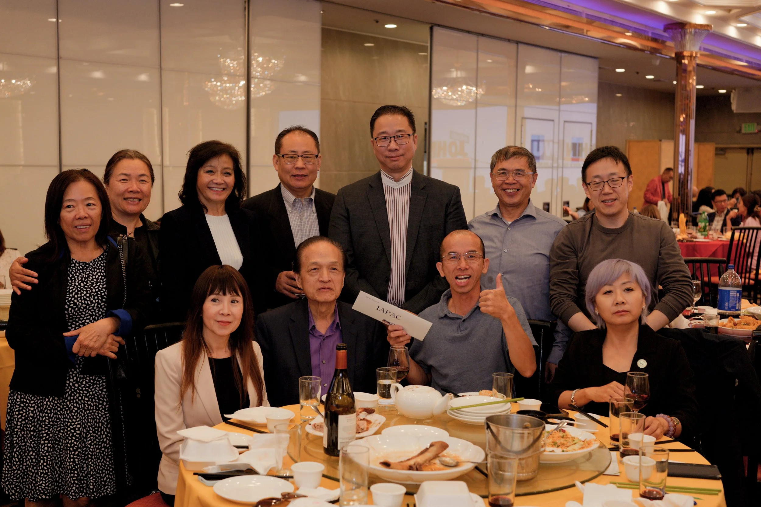 Group of people gathered around a banquet table at a formal event, with food and drinks, in an elegant hotel ballroom.
