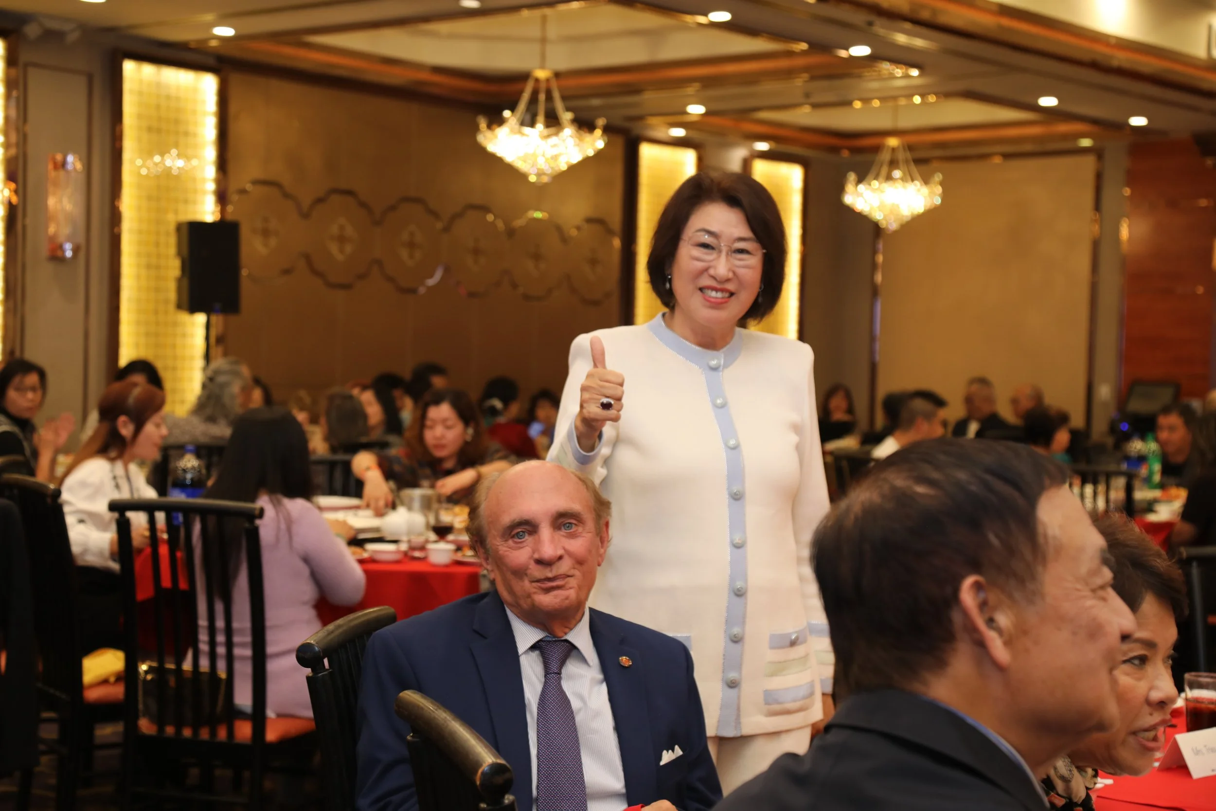 A woman in a white jacket giving a thumbs-up at a formal dinner event with a man in a suit seated in front of her, in a decorated banquet hall with chandeliers and other guests.