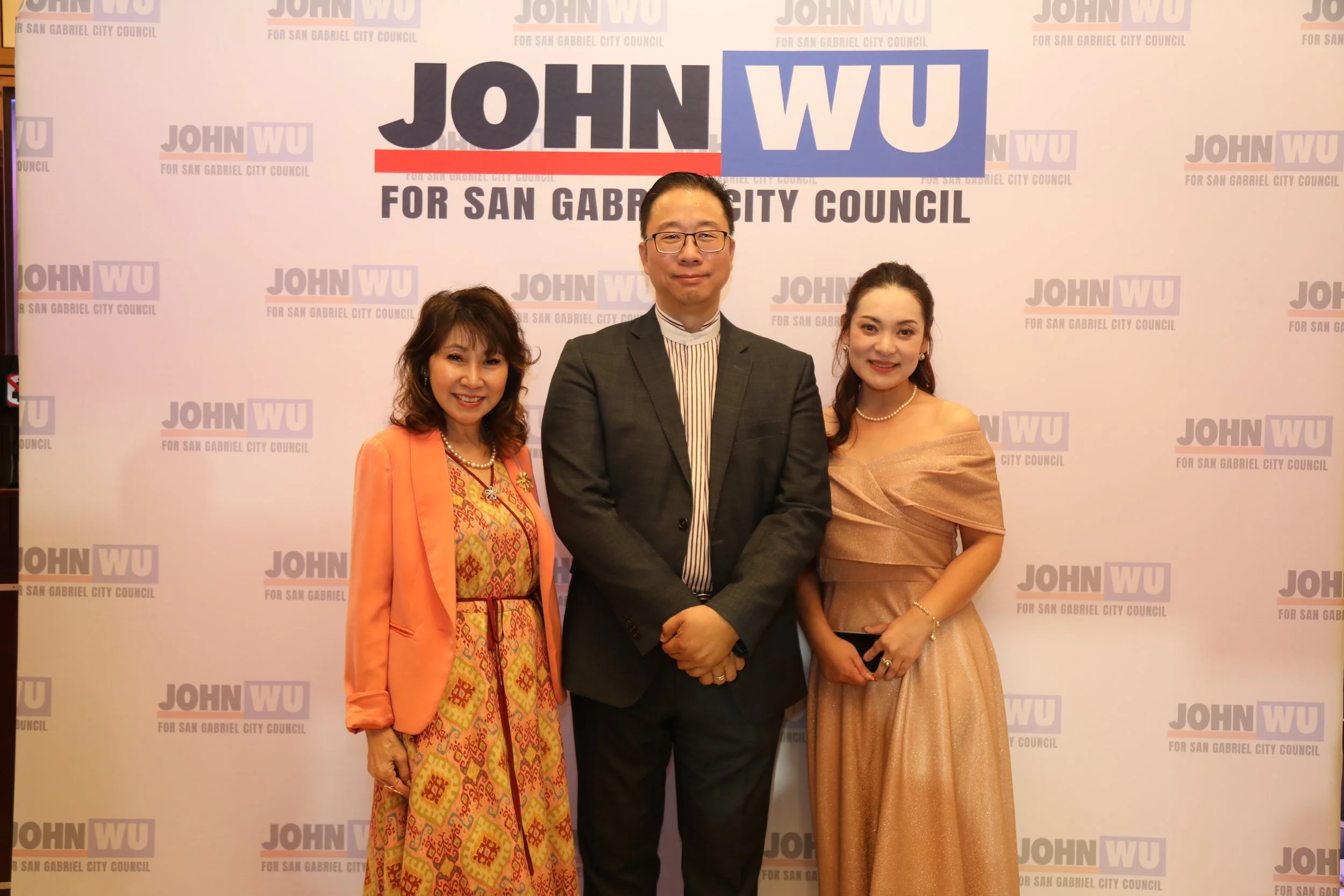 Three people standing in front of a backdrop with the text 'John WU for San Gabriel City Council.' The person on the left is a woman with brown hair wearing an orange blazer and a colorful patterned dress. The person in the center is a man with glass