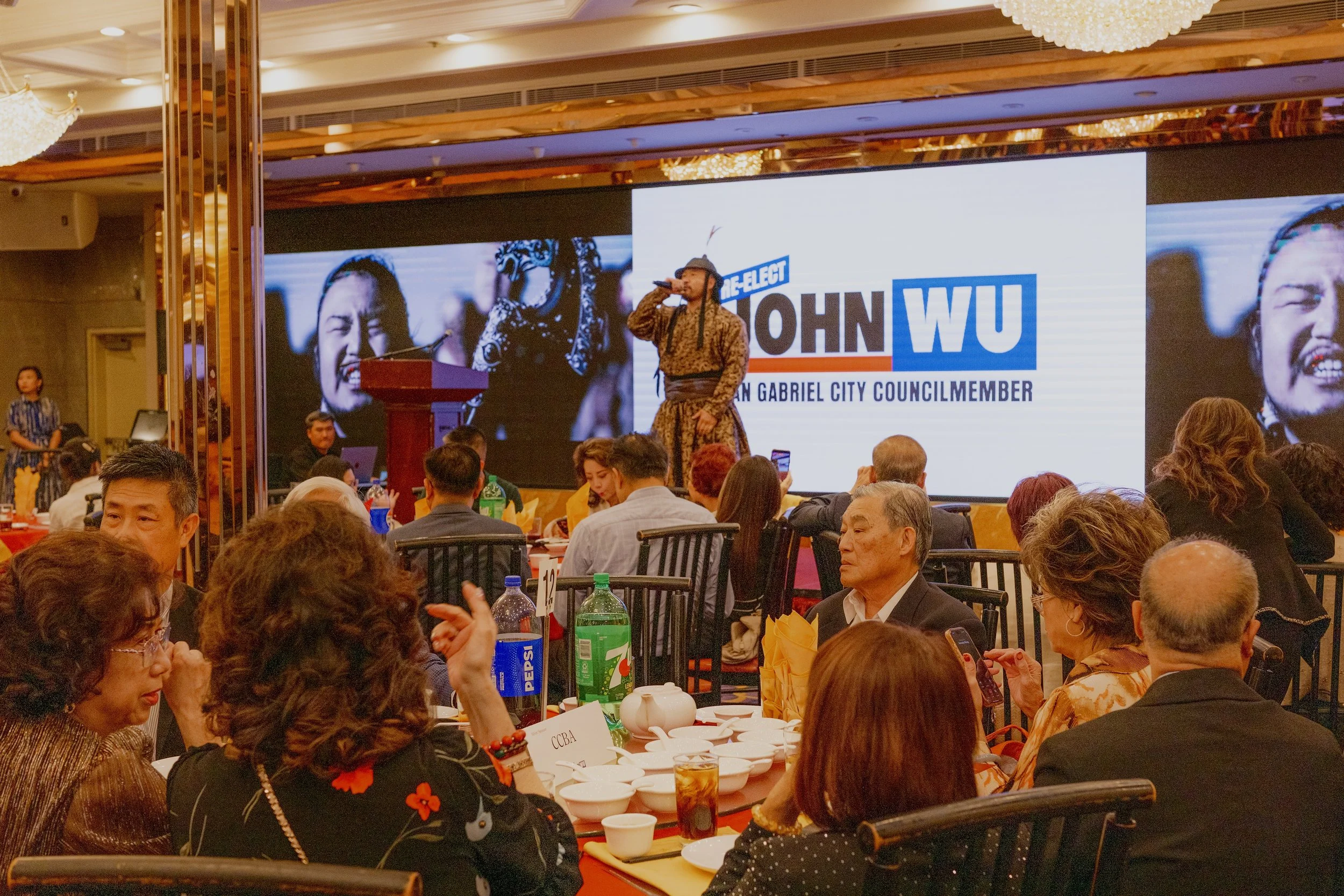 A man in traditional Filipino attire is speaking into a microphone on stage at a political event, with a large campaign sign behind him for John Wu, a candidate for GABRIEL City Council. The audience is seated at tables with food and drinks, and a wo