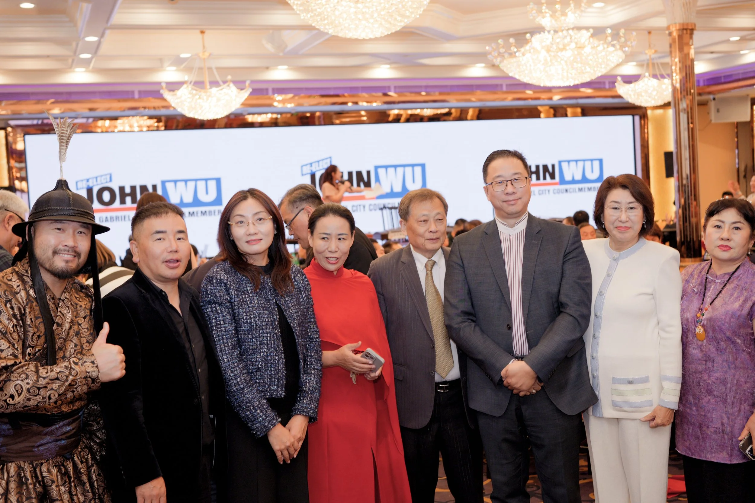 Group of diverse people posing at a formal event with a large screen in the background displaying 'John Wu' and 'City Council'. The room is decorated with chandeliers.