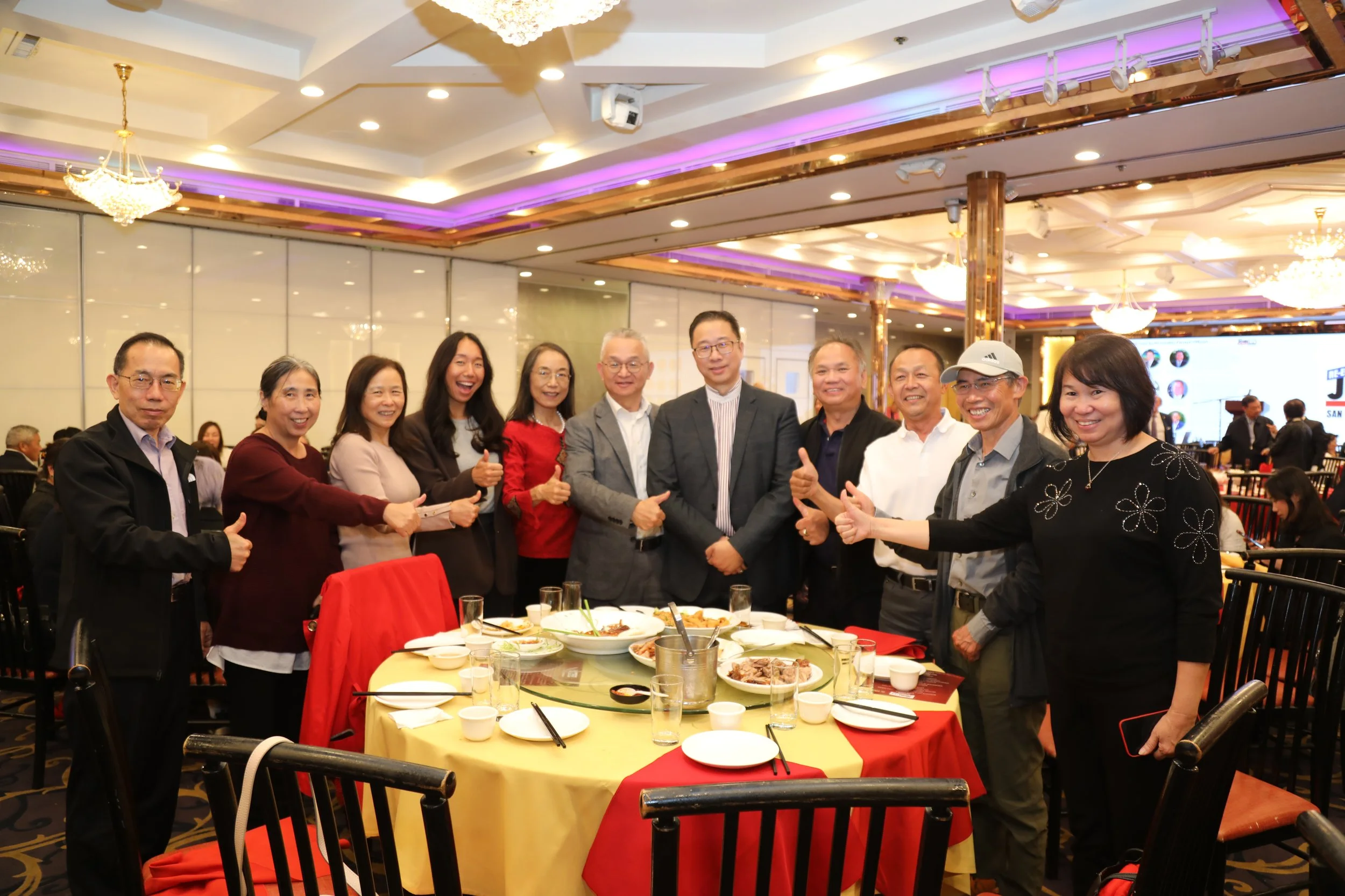 A group of diverse adults smiling and giving thumbs-up gestures around a banquet table with food in a decorated event hall with chandeliers and purple lighting.