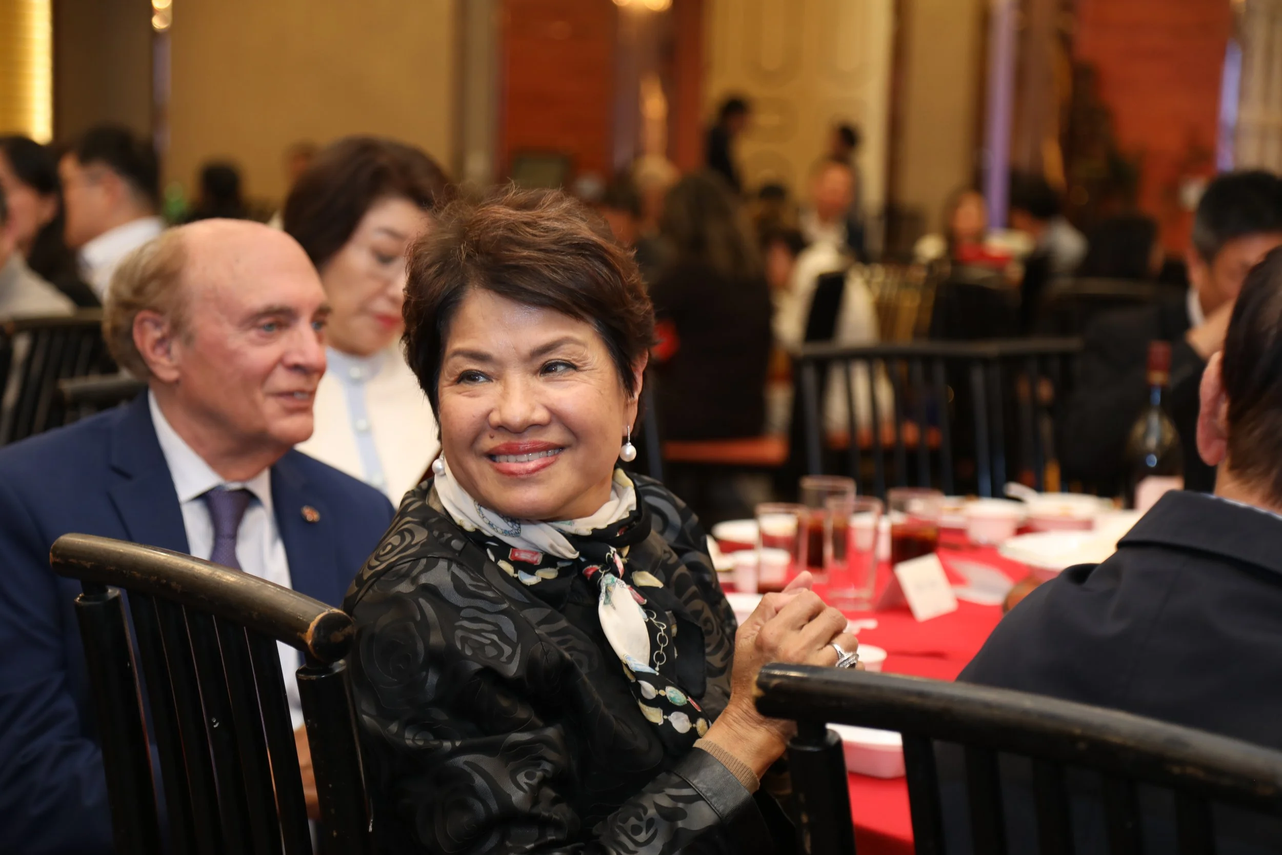 Various people attending a formal event, seated at round tables with red tablecloths, in an ornate ballroom, engaging in conversation and smiling.