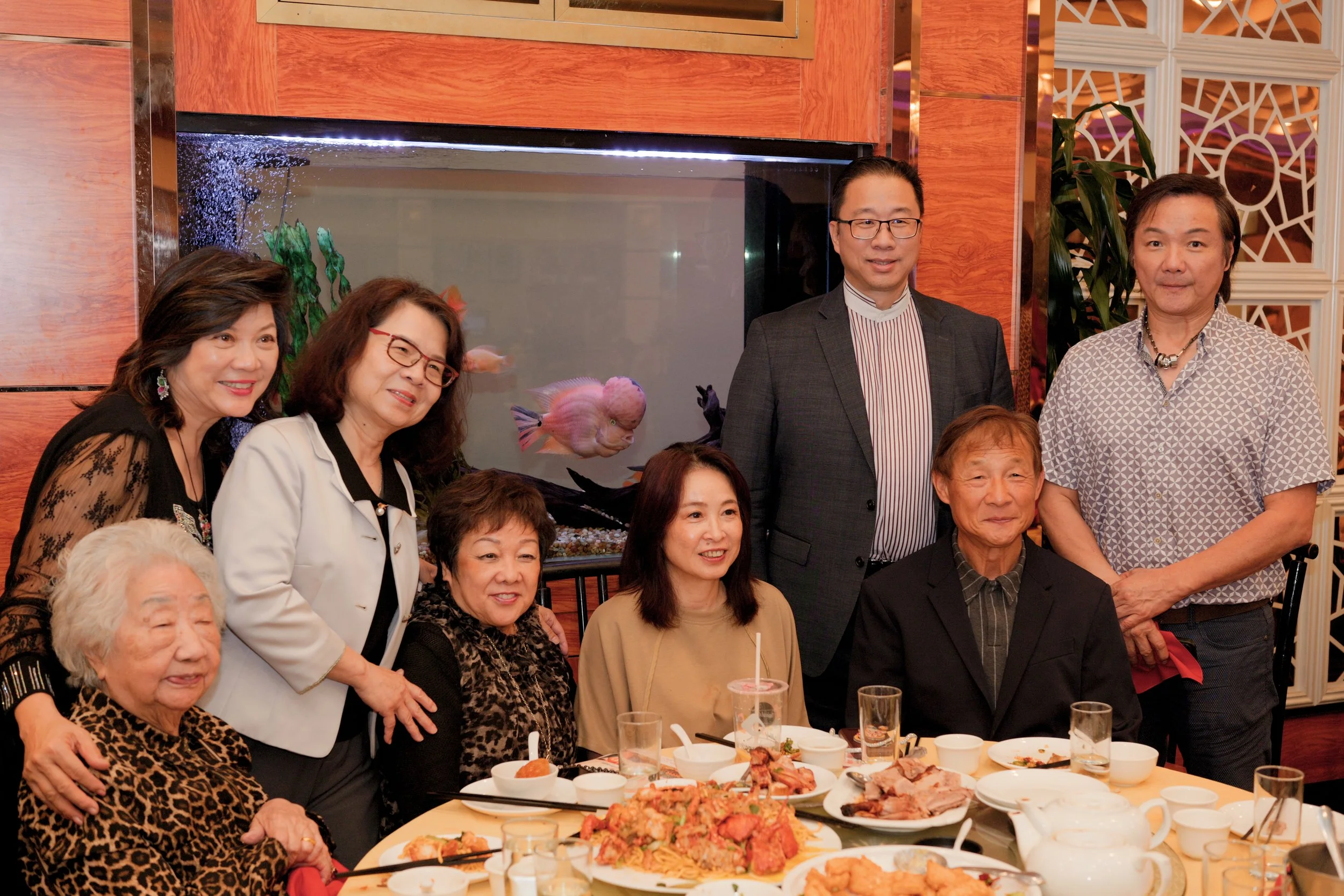 A group of diverse people gathered around a banquet table with food, posing for a group photo in a restaurant. Fish tank visible in the background.