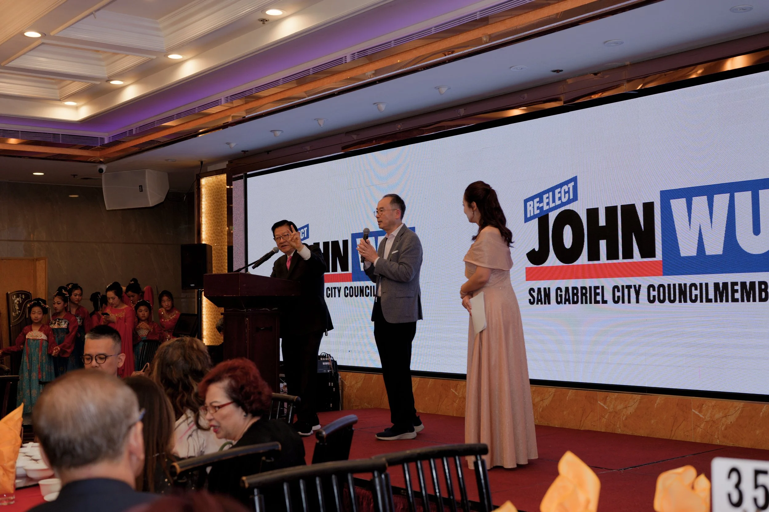 A political event with a stage, large screen displaying a campaign sign for John Wu, San Gabriel City Councilmember, and several people on stage including a man speaking into a microphone, a woman in a peach dress, and another man in a gray suit. The