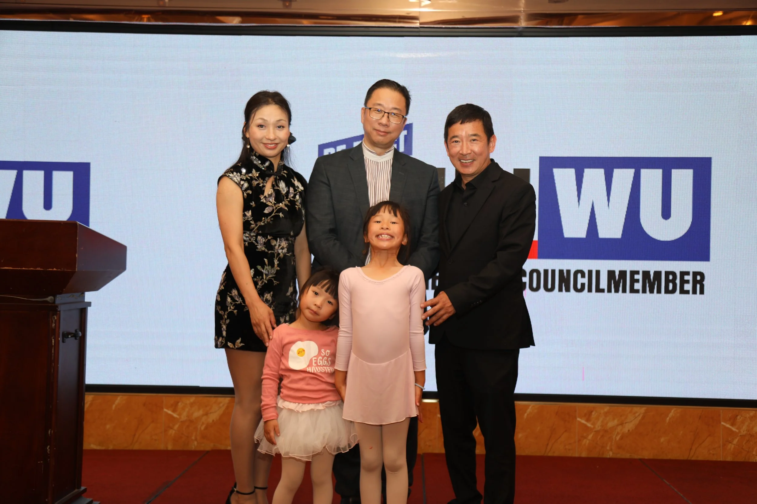 A group of three adults and three children standing together at an event with a large screen in the background displaying the words 'REPUBLIC' and 'COUNCILMEMBER'. The woman on the left is wearing a black floral dress, the man in the middle is in a s