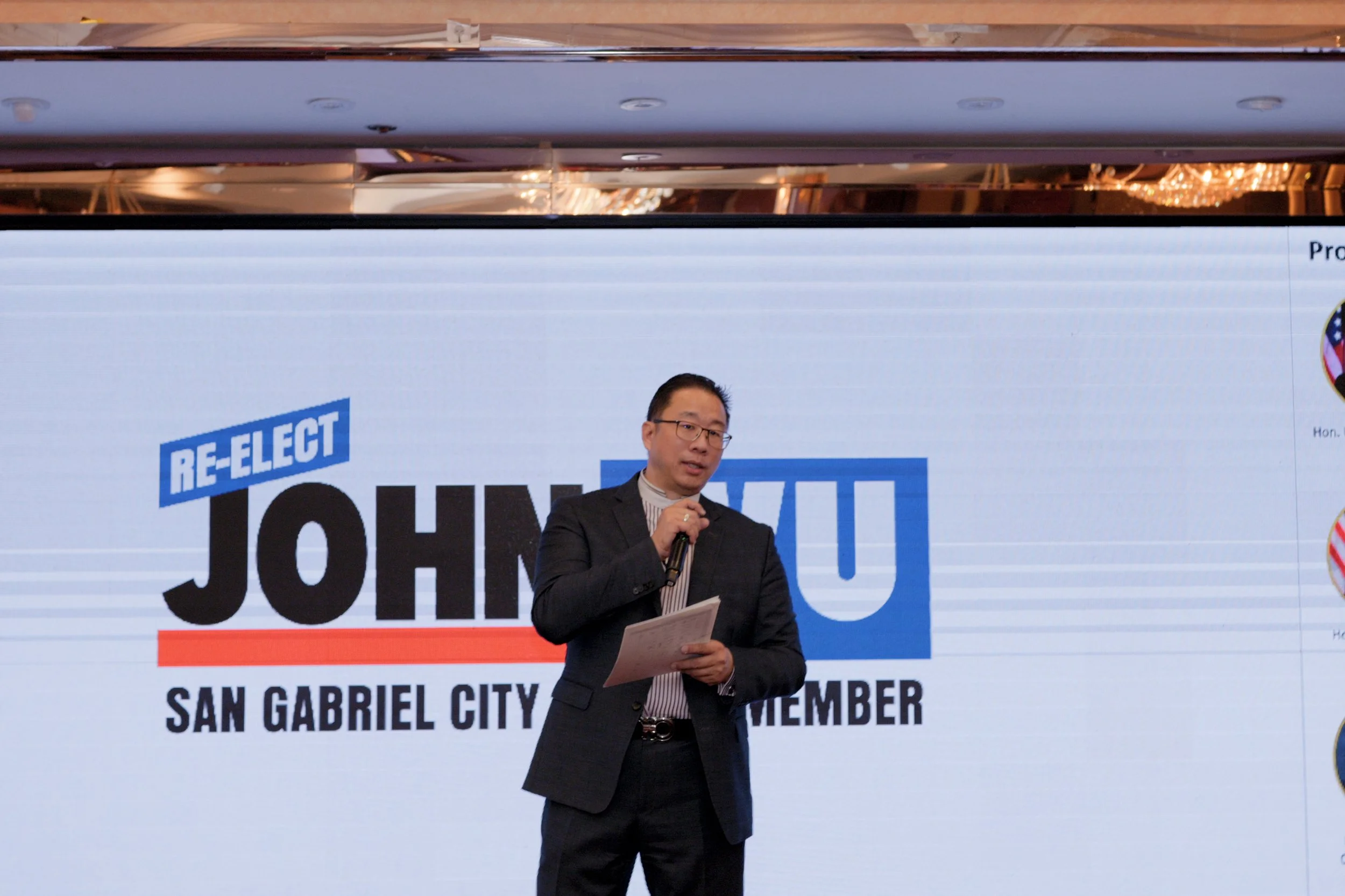 A man in a suit and glasses speaking into a microphone at a political campaign event. Behind him is a large display with the text "Re-Elect" and "JUNE 7" along with the name "John" and location "San Gabriel City".