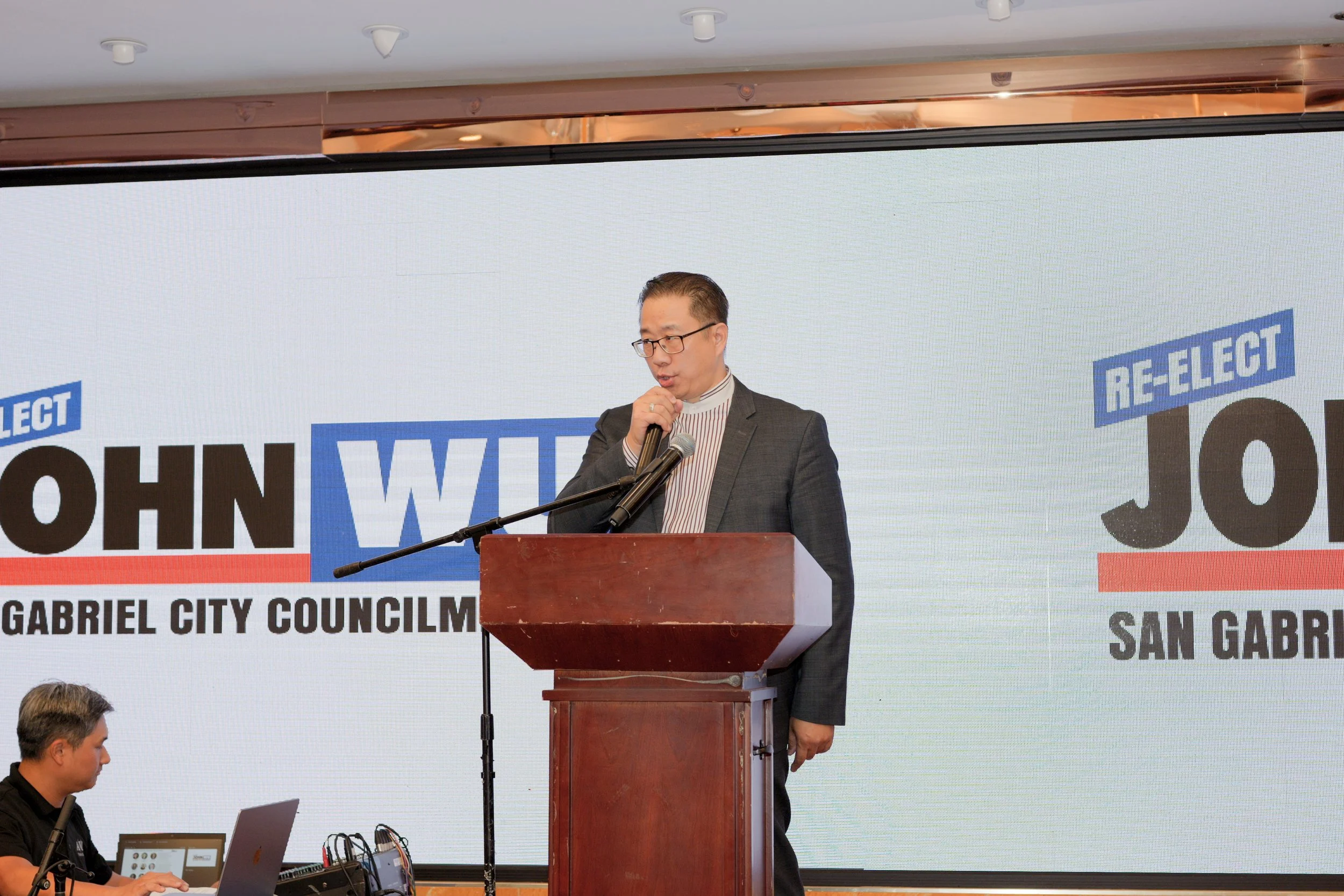 A man in a gray suit and glasses is speaking into a microphone at a podium during a public event. Behind him is a large banner with text indicating the event is related to John Wu for San Gabriel City Council. A man on a laptop is seated in front of 