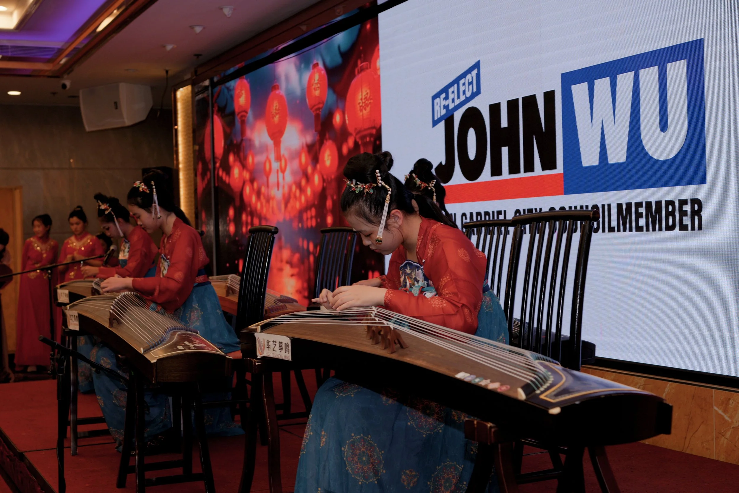A group of women in traditional red and blue dresses playing the guzheng, a Chinese musical instrument, on a stage during an event. Large screen behind them displays the text 'RE-ELECT JOHN WOU' and other partial text.