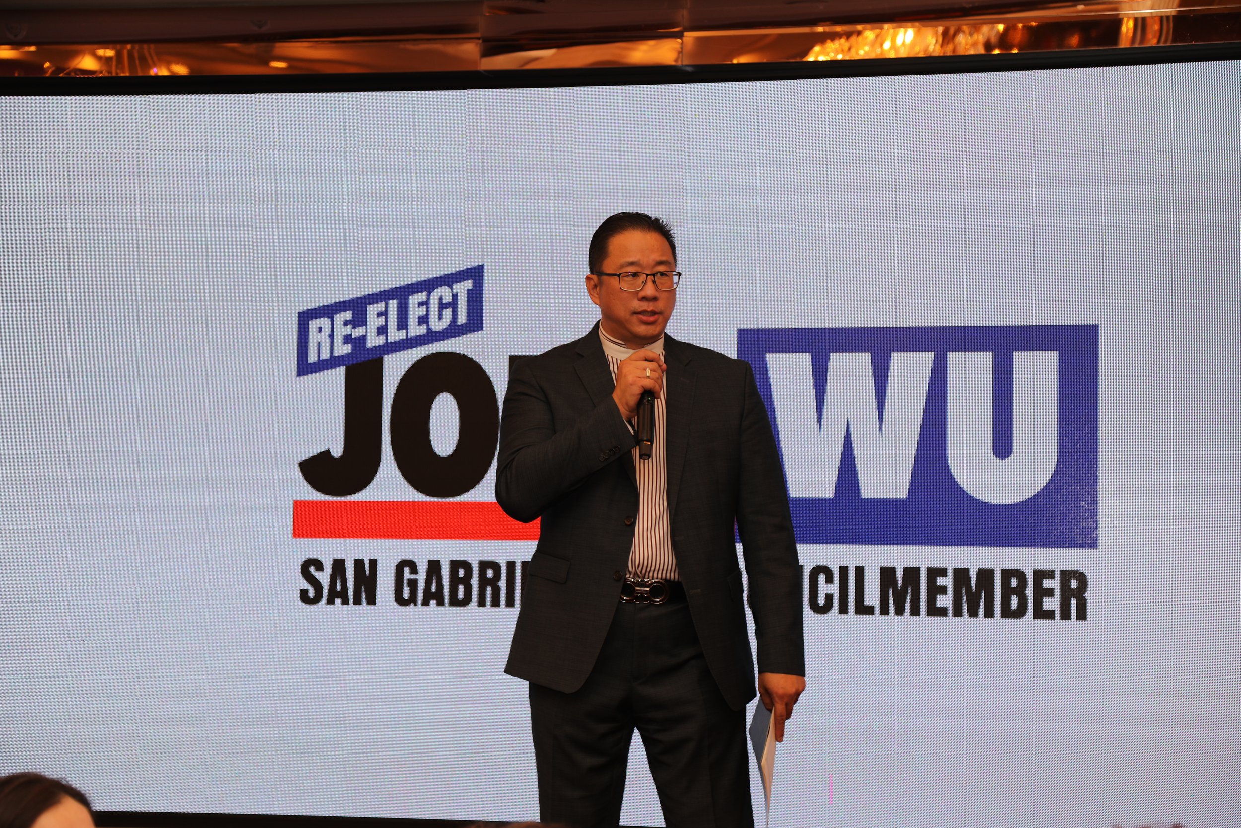 A man in a suit and tie holding a microphone, standing in front of a large digital screen displaying text 'RE-ELECT' and 'JOW WU' with additional smaller text 'SAN GABRIEL CITIES COUNCILMEMBER'