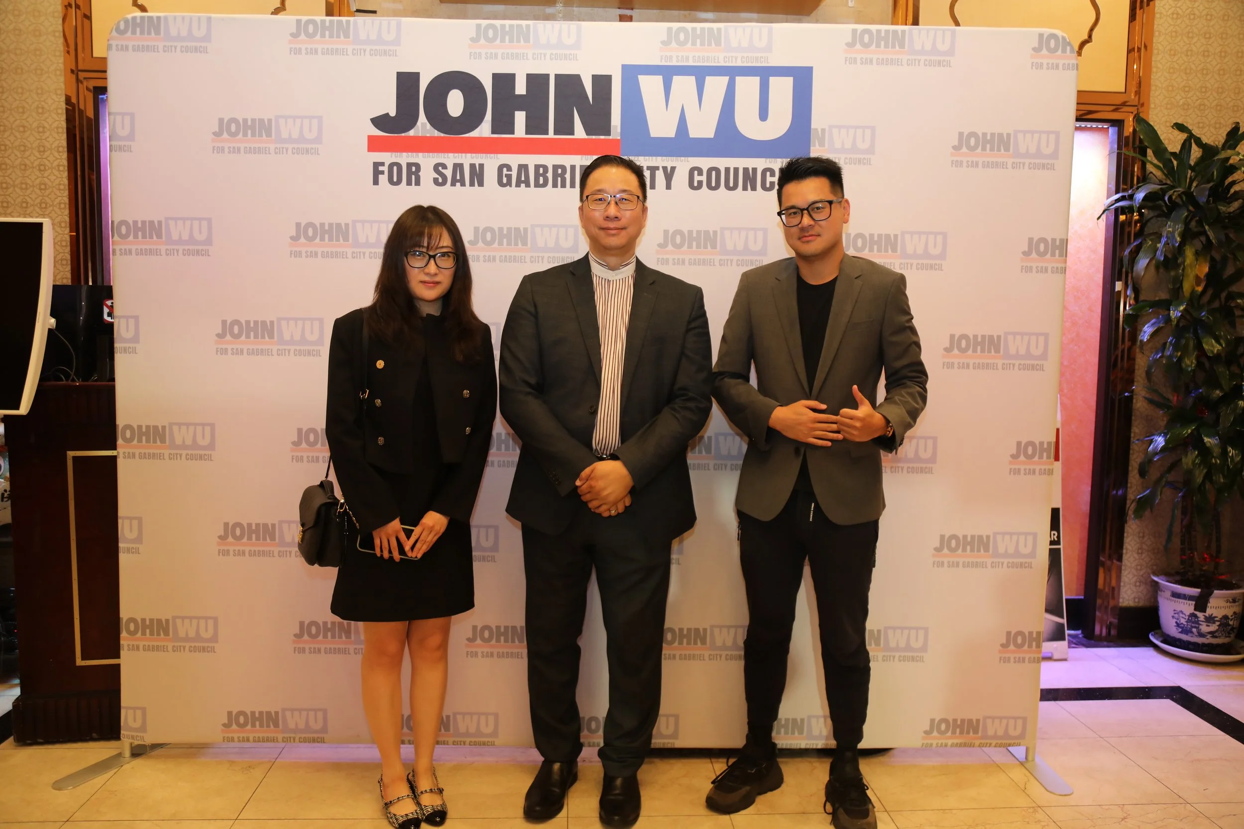 Three people standing in front of a campaign backdrop featuring the name 'John Wu' and the phrase 'For San Gabriel City Council'. The group includes a woman on the left wearing glasses and a black blazer, a man in the middle with glasses, a suit, and