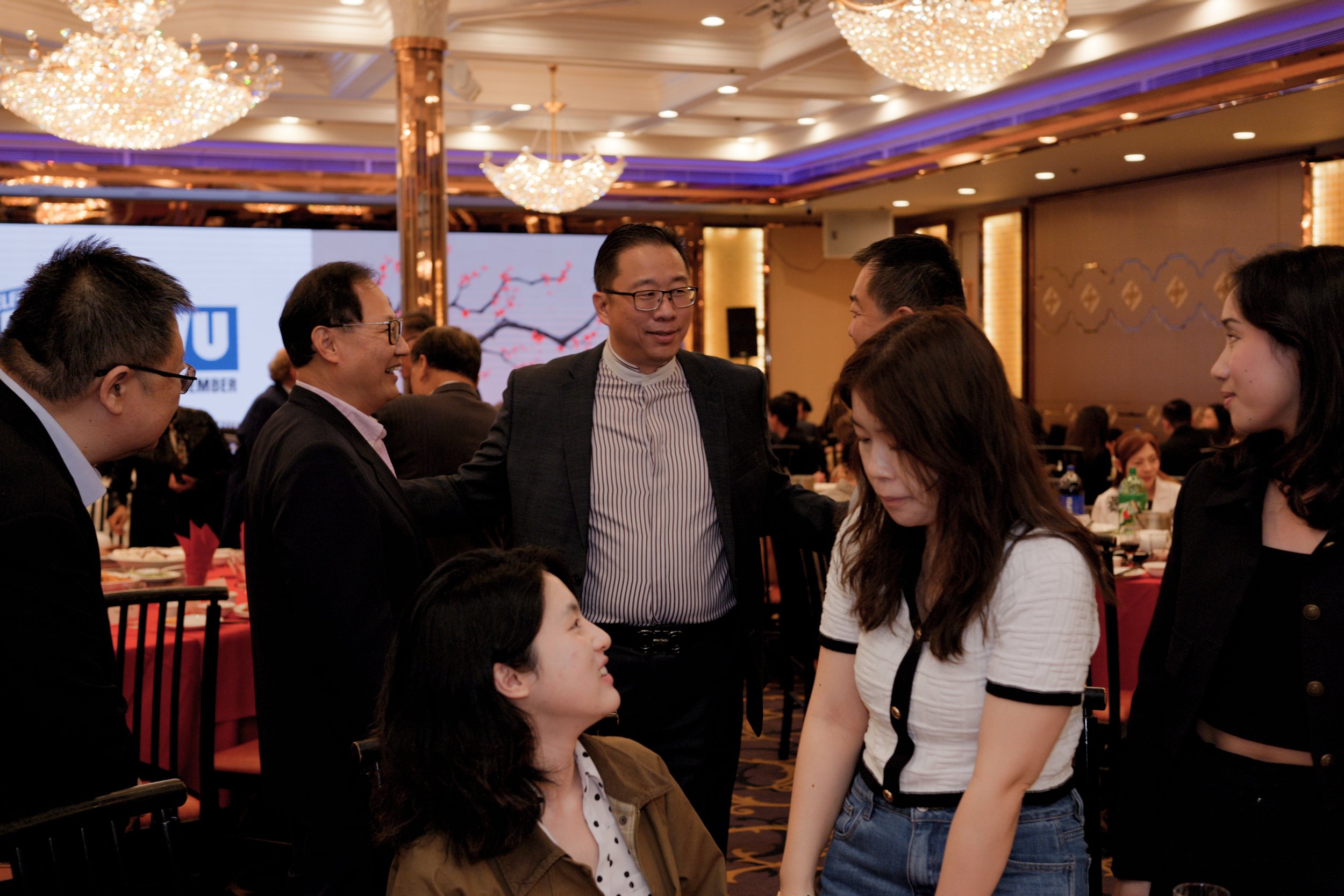 Group of people engaging in conversation at a formal event or conference, with ornate chandeliers and decorative lighting in the background.