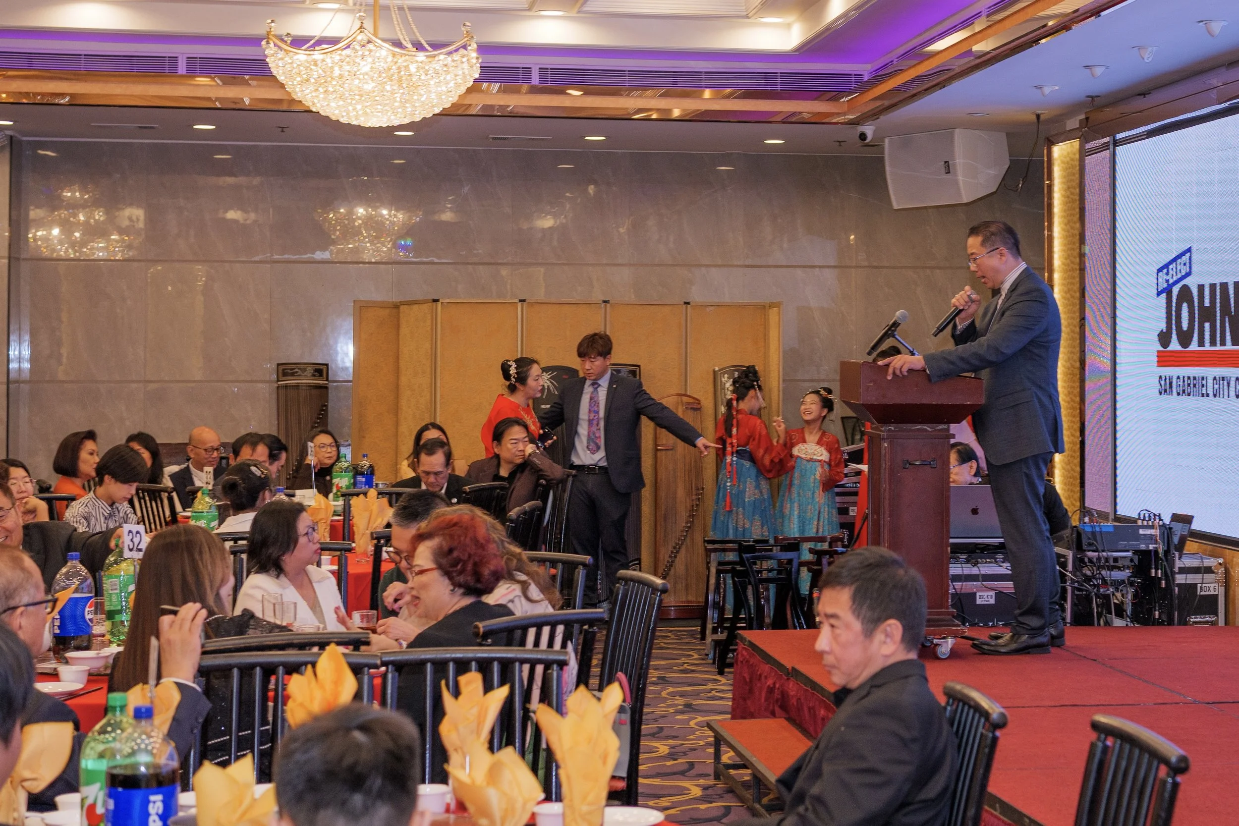 A man in a suit giving a speech at a podium with a microphone at an event. In the background, there are people dressed in traditional Asian clothing, and an audience seated at tables with beverages and napkins. The setting appears to be a banquet hal