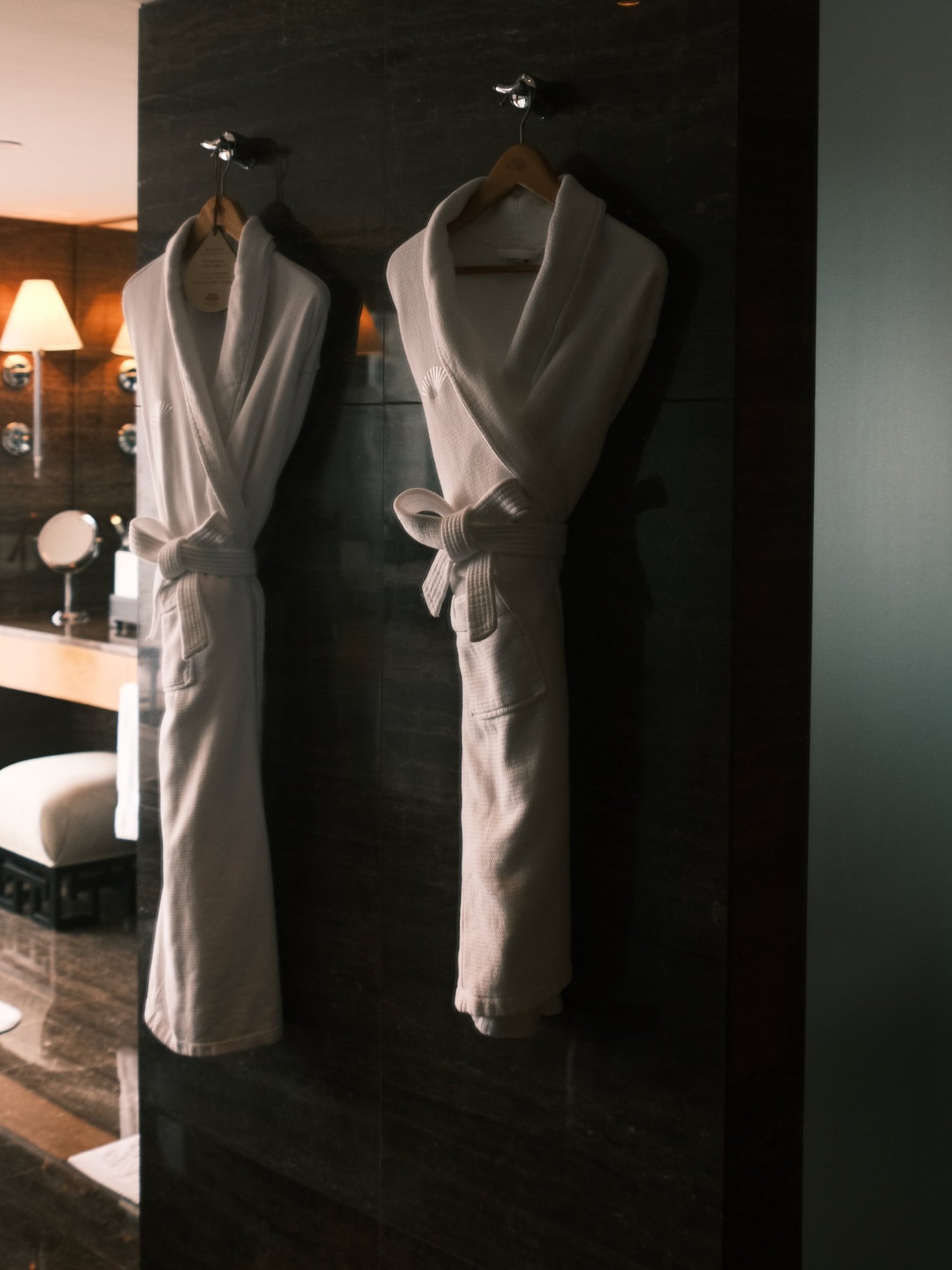 Two white bathrobes hanging on hooks on a dark wall inside a hotel or spa room.