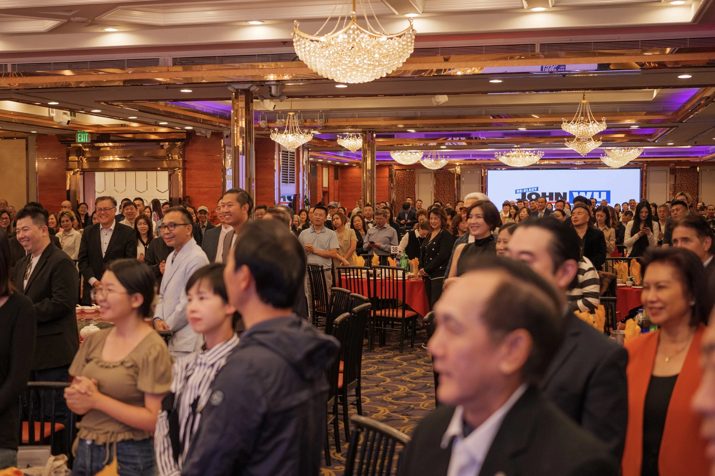 A large group of people attending a conference or event in a banquet hall with elegant chandeliers and purple lighting. The audience is smiling and engaged, with some taking photos. A screen in the background displays a political campaign logo for Jo