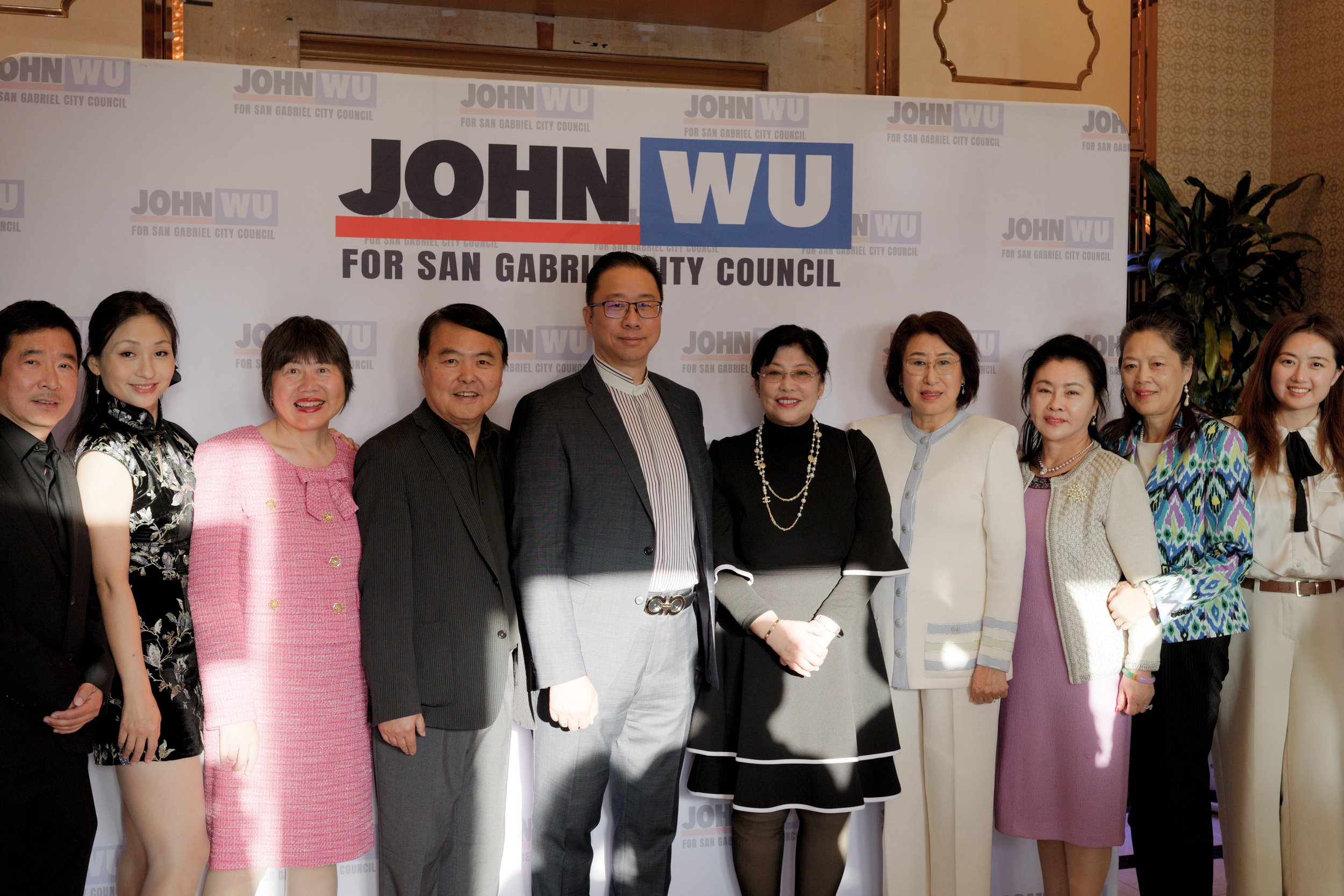A group of eleven people standing together in front of a campaign banner that reads 'JOHN WU FOR SAN GABRIEL CITY COUNCIL.' They are dressed formally, smiling, and posing for the photo in an indoor setting with a decorative background and potted plan