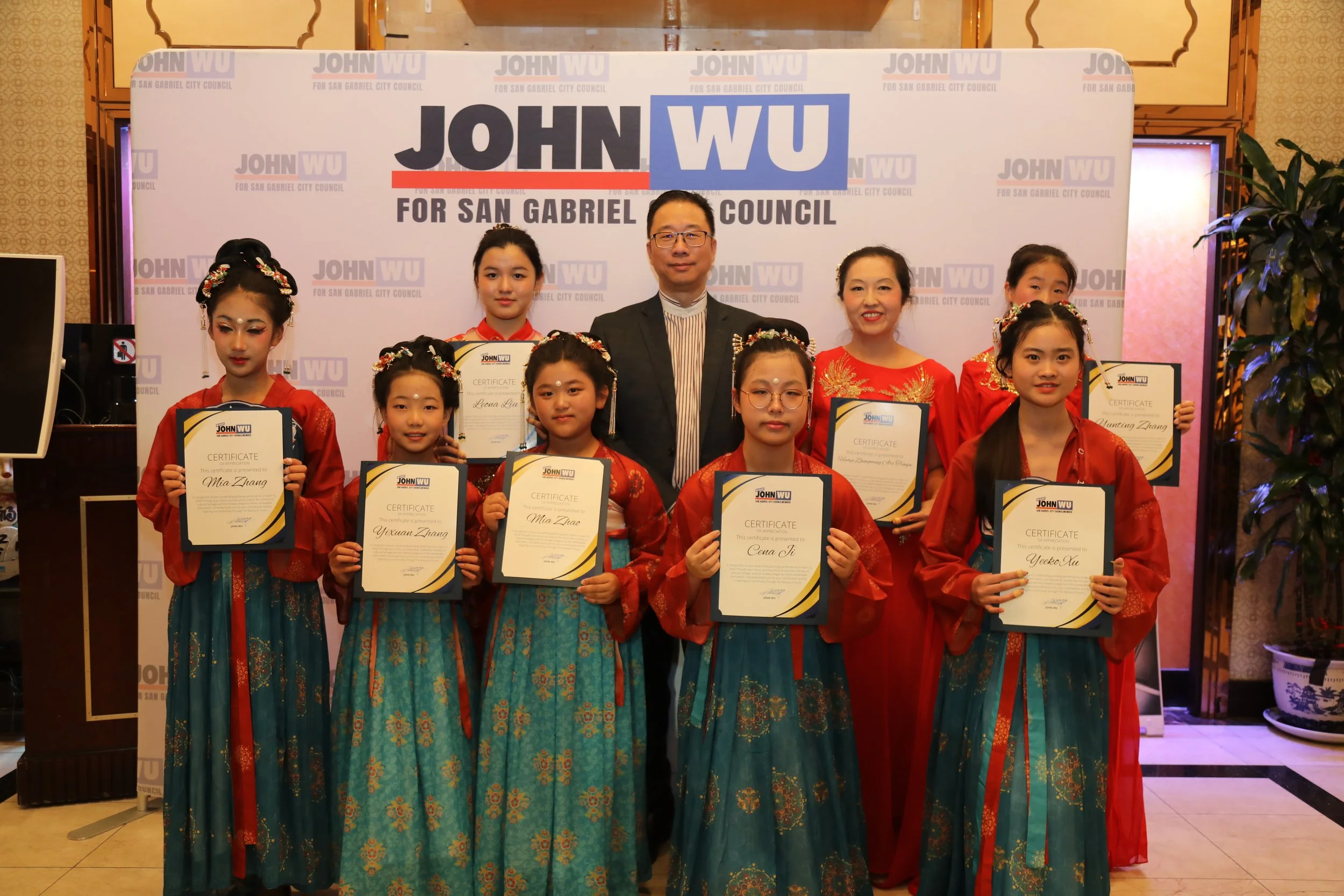 Group of young girls in traditional Chinese dresses holding certificates, standing with two adults, in front of a backdrop that reads 'John Wu for San Gabriel City Council'.