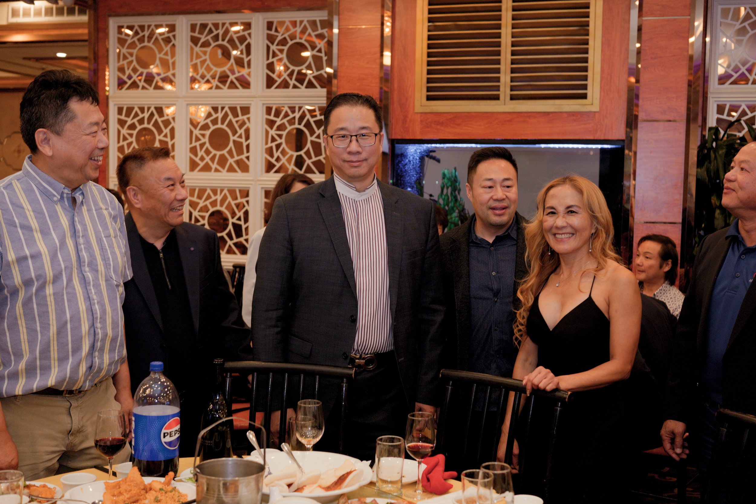 Group of people at a dinner party in an elegant setting, smiling and posing for a photo, with food and drinks on the table.
