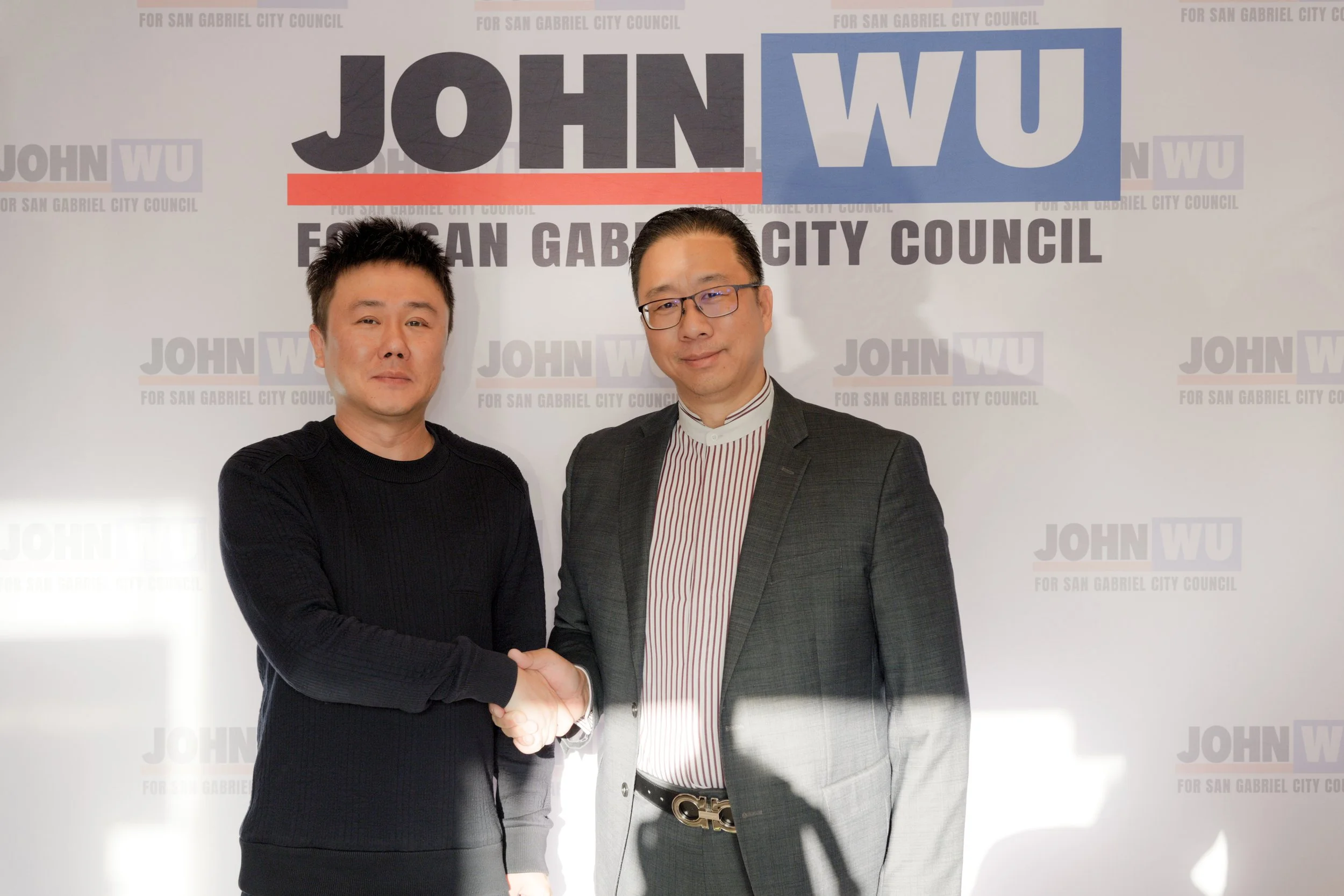 Two men shaking hands in front of a campaign sign for John Wu for San Gabriel City Council. The man on the left is wearing a black sweater, and the man on the right is wearing glasses, a striped dress shirt, and a suit jacket.