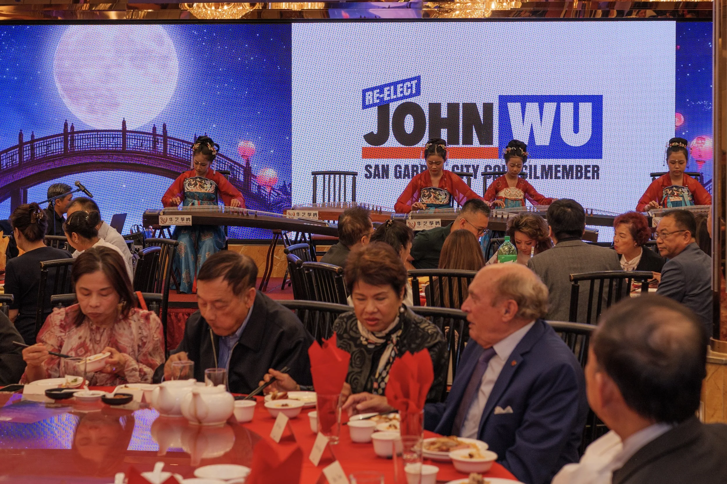 People dining at a banquet with a large screen in the background displaying the name and photo of John Wu, a city council member from San Gabriel.