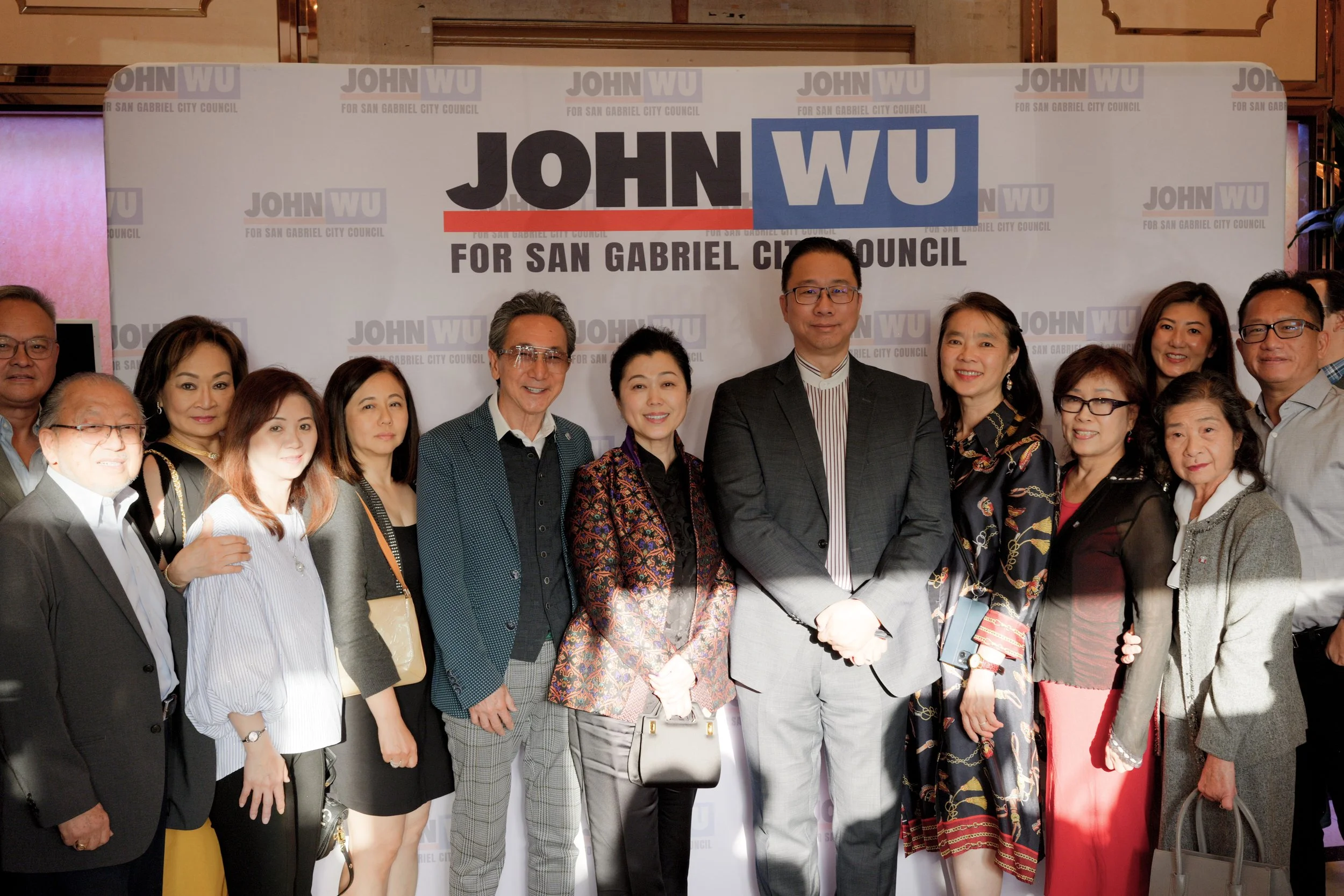 Group of people standing together at a political campaign event for John Wu for San Gabriel City Council, with a backdrop displaying the candidate's name and campaign slogan.