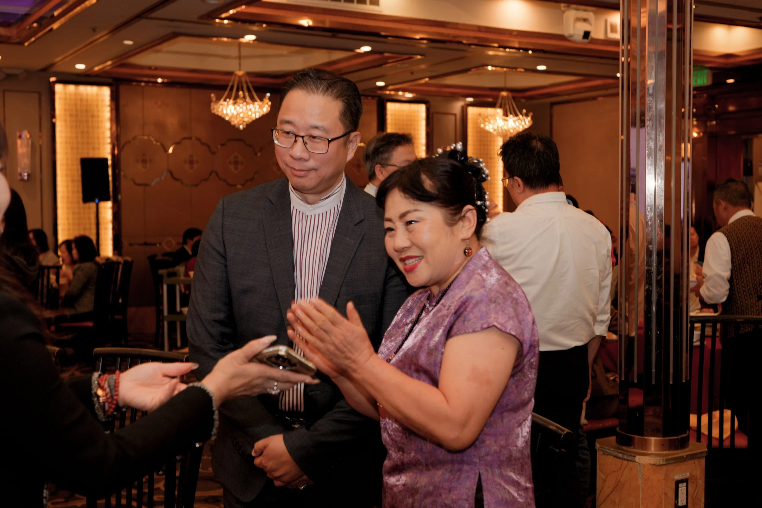 A man wearing glasses and a gray suit interacting with a woman in a pink patterned dress at a formal event, with other people and elegant chandeliers in the background.