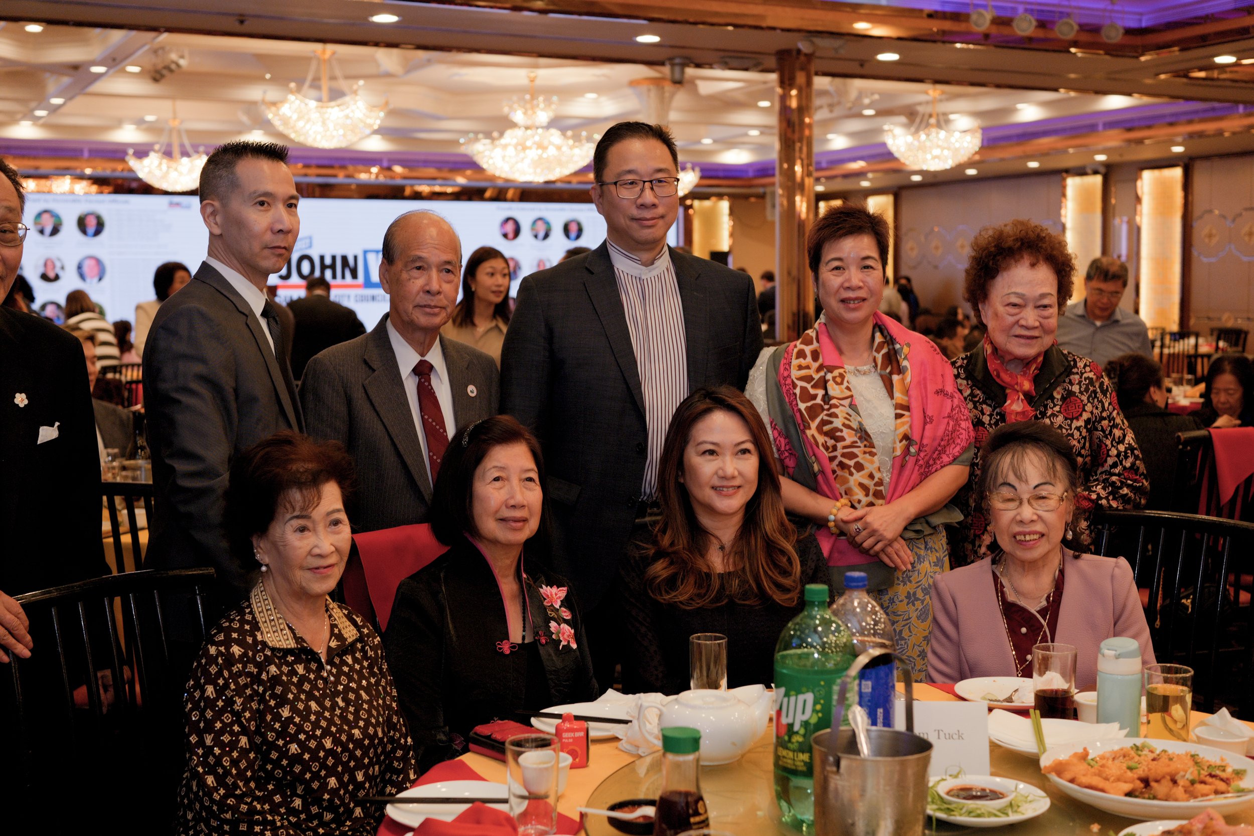 Group of people, mostly Asian women and men, dressed in formal attire, standing and sitting at a banquet table in a decorated ballroom with chandeliers, during a social or formal event.