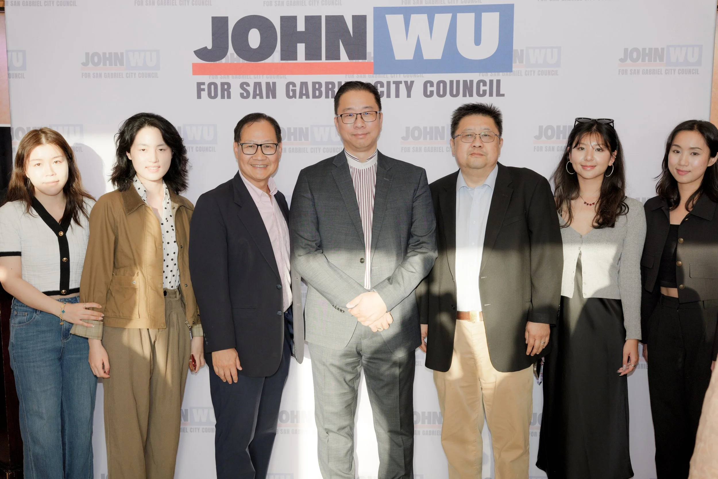 A group of seven people, four women and three men, standing together in front of a backdrop with political campaign text. The backdrop reads: 'JOHN WU FOR SAN GABRIEL CITY COUNCIL.' The individuals are dressed in professional attire, and three of the