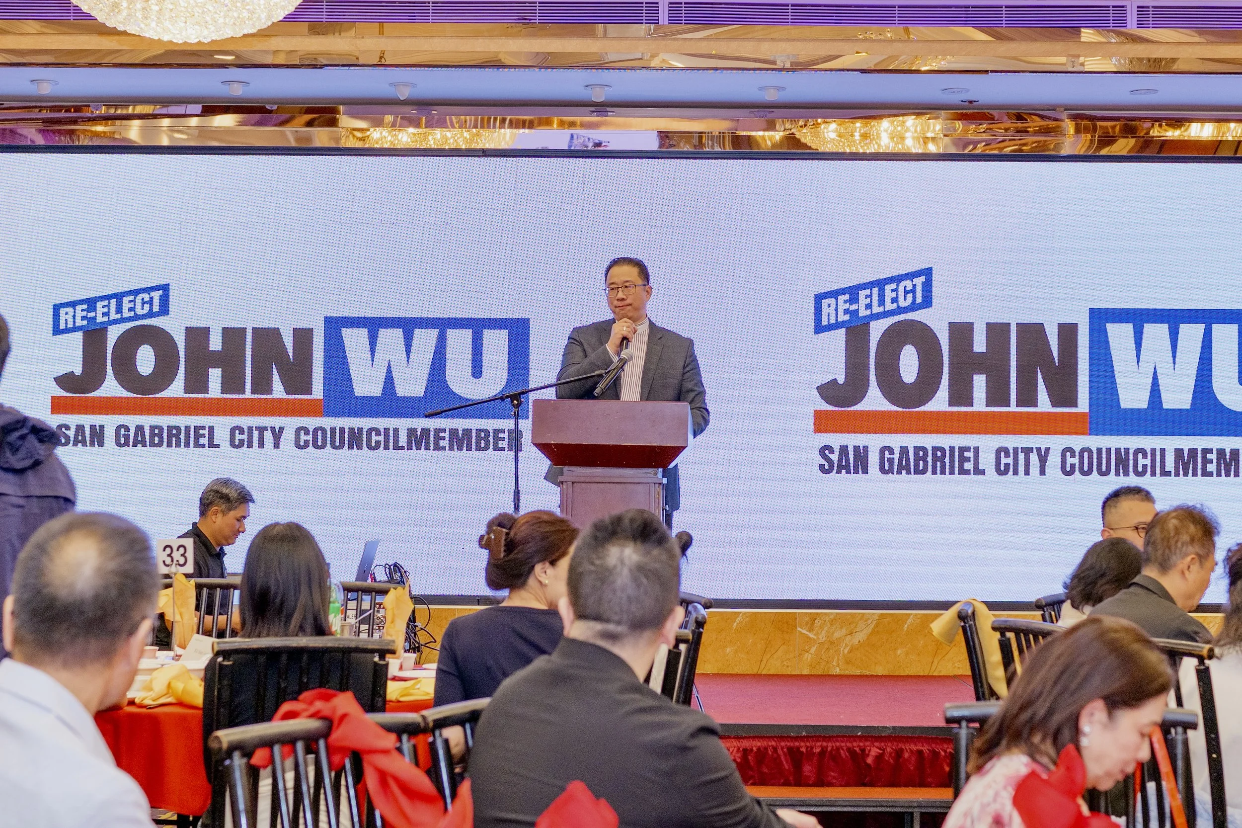 A man in a suit standing at a podium, speaking into a microphone, in front of large digital display screens with campaign signs for John Wuo, a candidate for San Gabriel City Council Member. The audience is seated at tables with people and items, lis