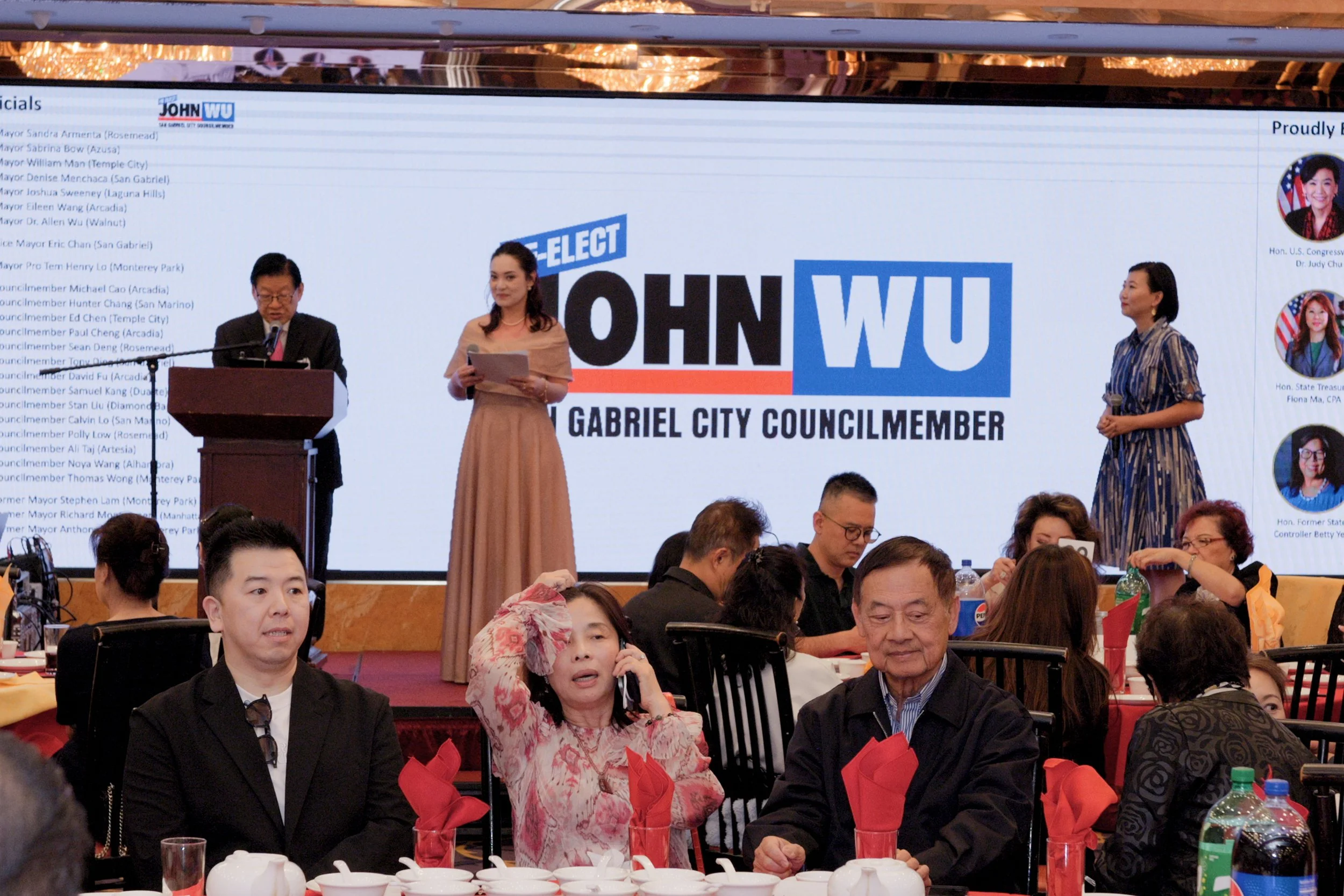 A group of people seated at a banquet table with red napkins, some eating and drinking, attending an event. In the background, there is a large screen displaying the name 'JOHN WU,' and a woman standing in front of it, dressed in a beige dress, holdi