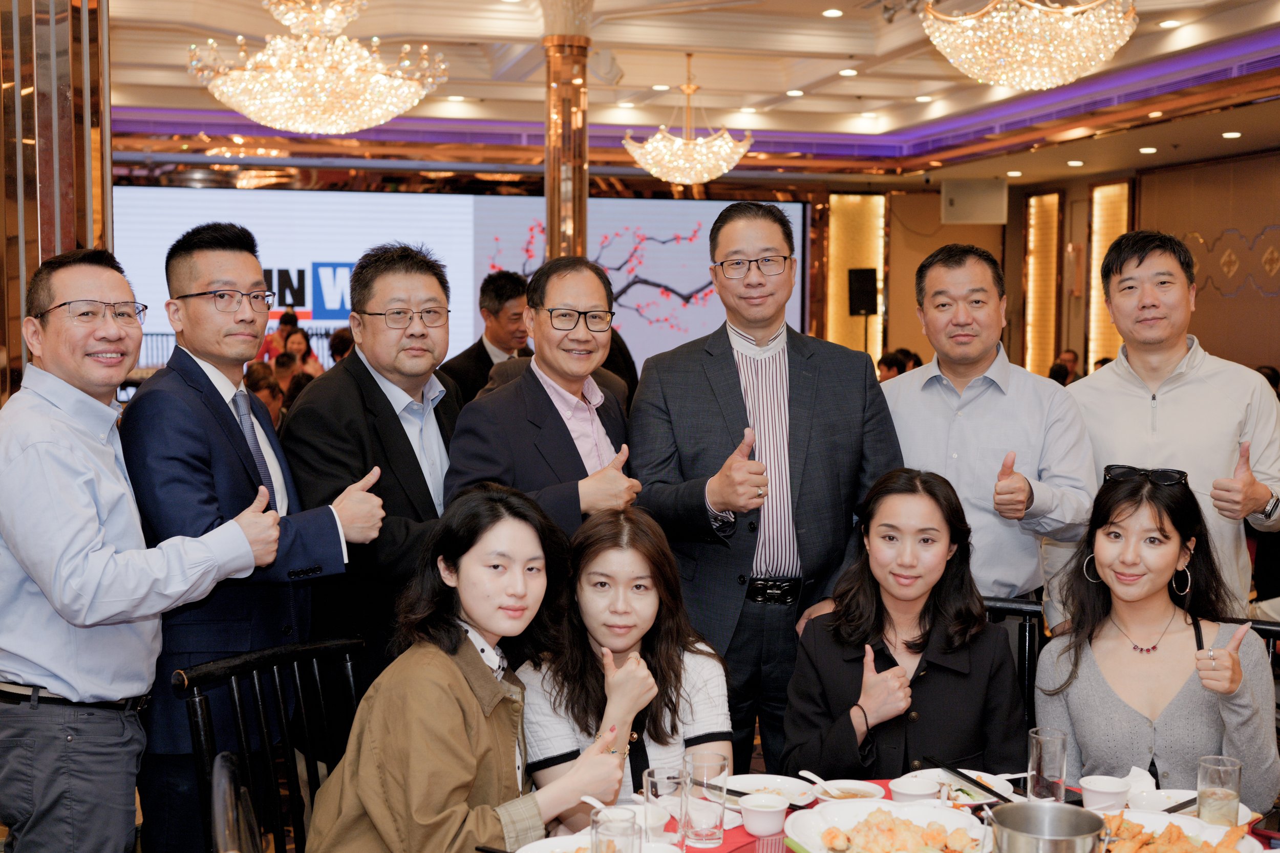 Group of Asian professionals and women at a formal event or celebration in a banquet hall with elegant chandeliers, a large digital screen, and tables set with food and drinks.