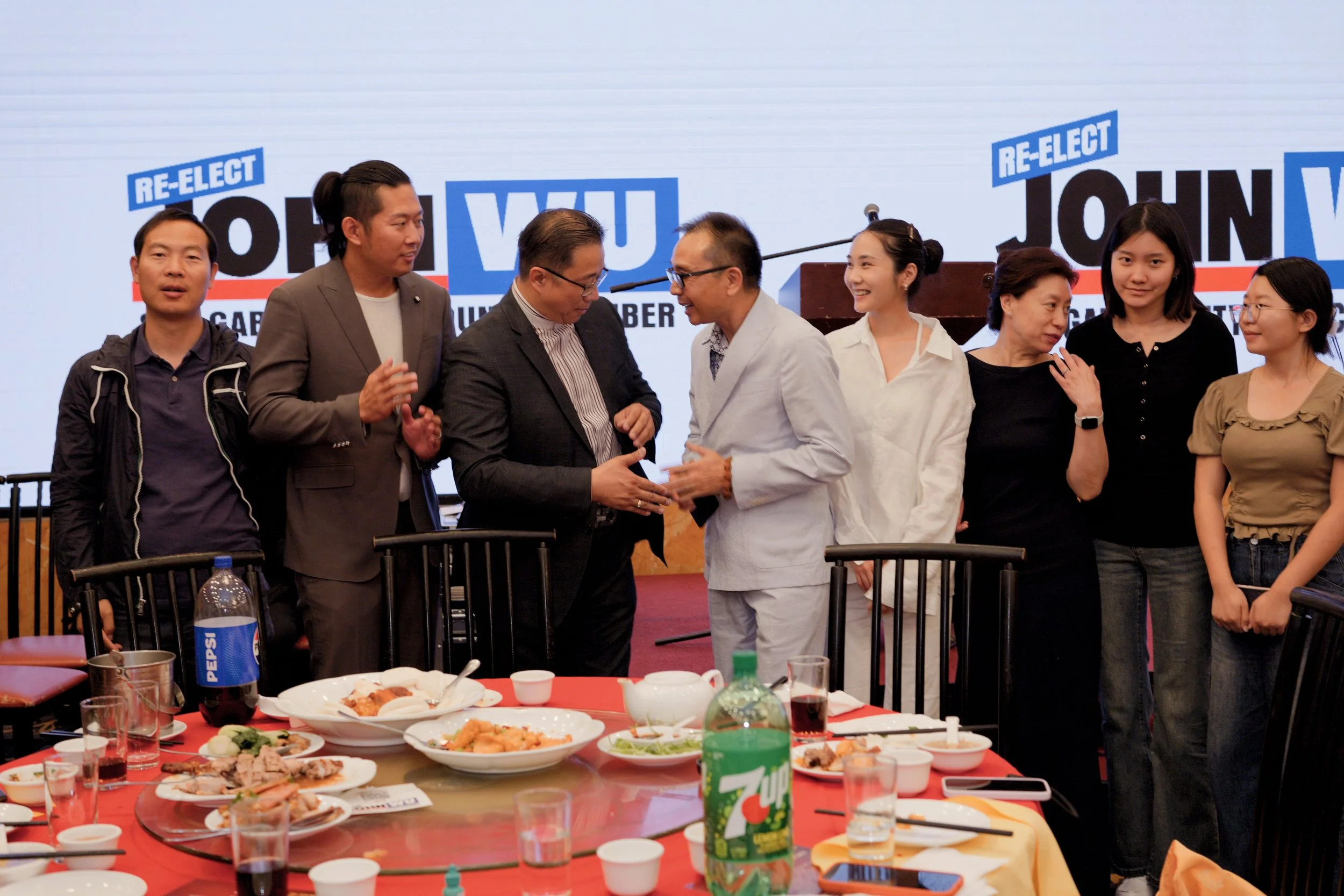 Group of people at a meeting or celebration with food, drinks, and a large screen in the background displaying a slogan, re-elect John W. Y. campaign.