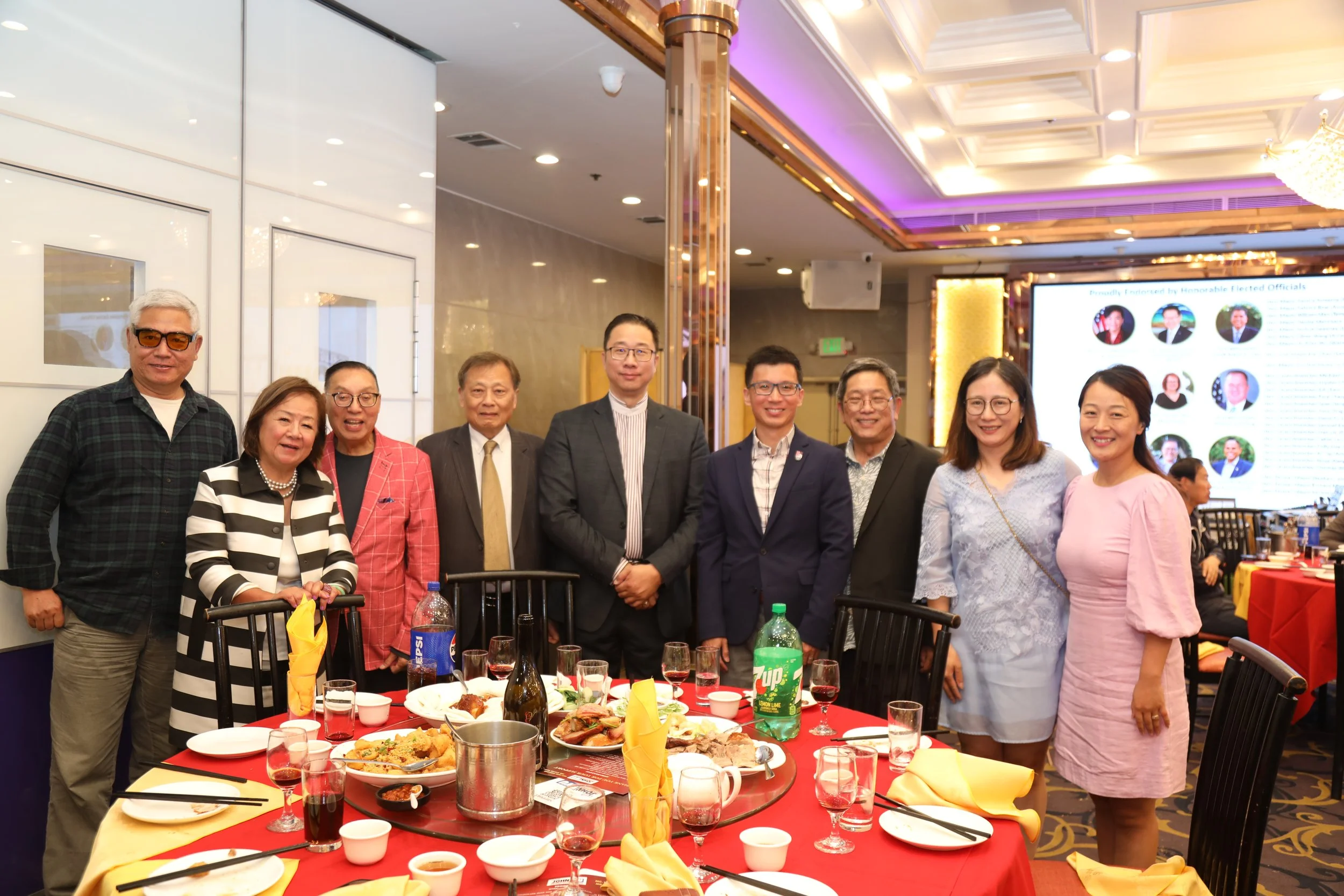A group of people standing together at a banquet table in a decorated event hall. There are various dishes and drinks on the table, and a large screen displaying images and text in the background.