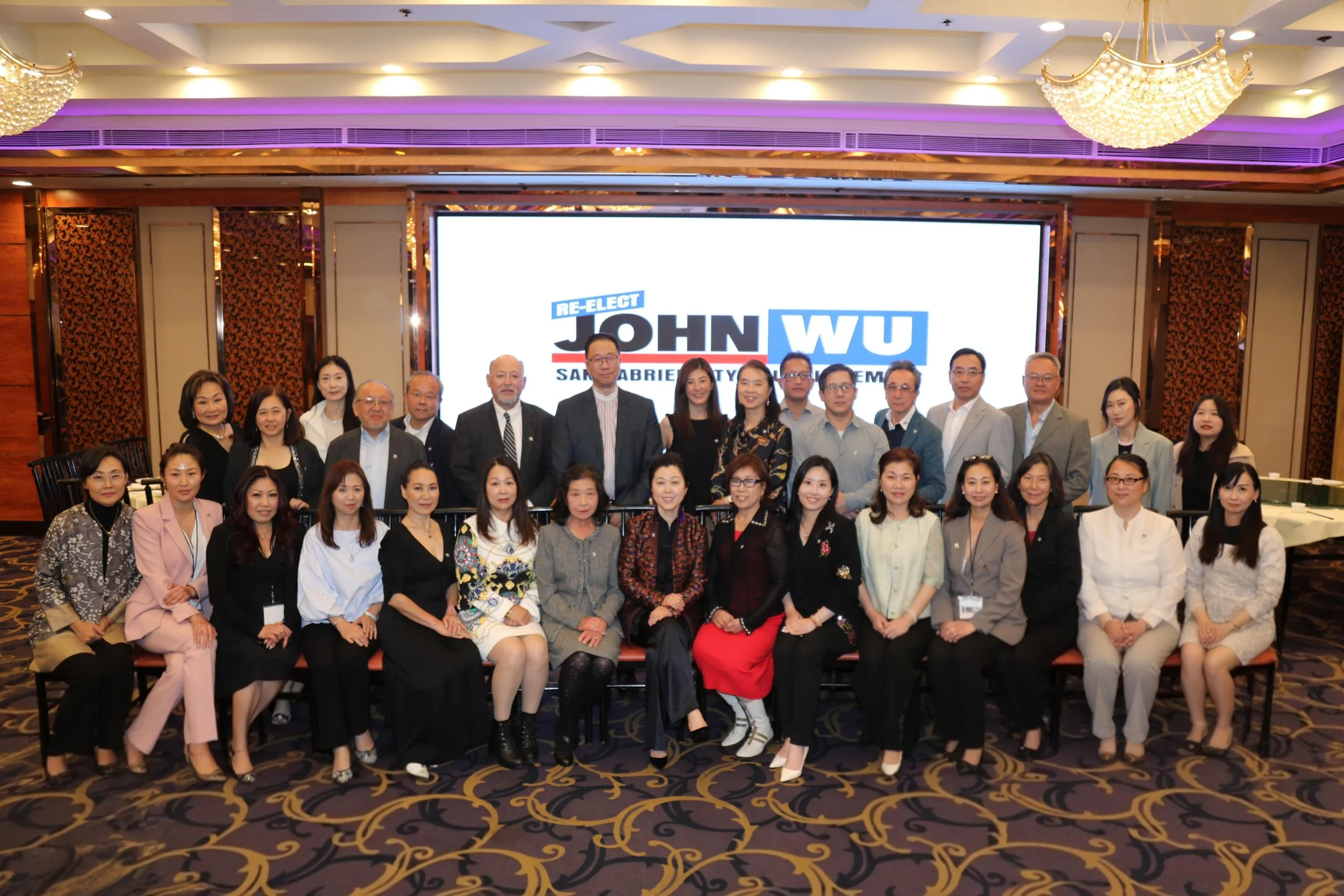 Group of people in formal attire posing for a photo at an event with a large screen displaying political campaign logos in the background.