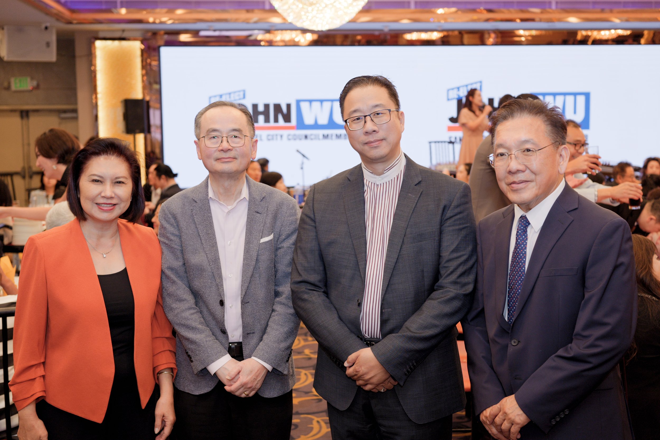 Four people standing together at a formal event, with a large screen in the background showing the names of officials involved in a local city council meeting.