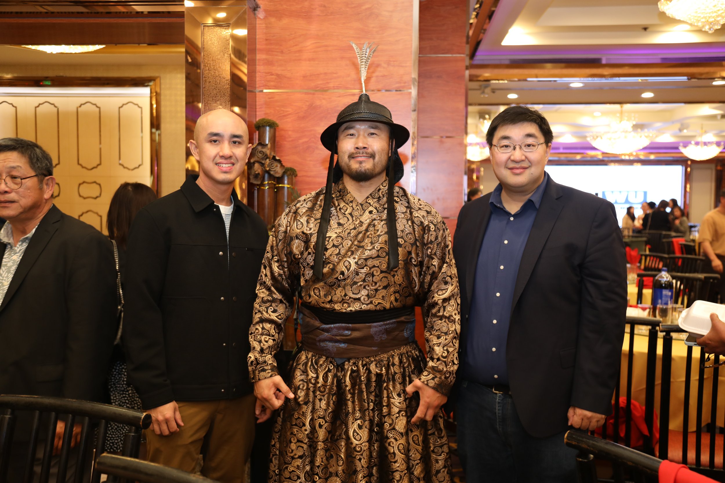 Four men standing together at an event in a ballroom with ornate decor and chandeliers. One man wears traditional Asian attire with a feathered hat, while the others are dressed in modern business casual and formal clothing.