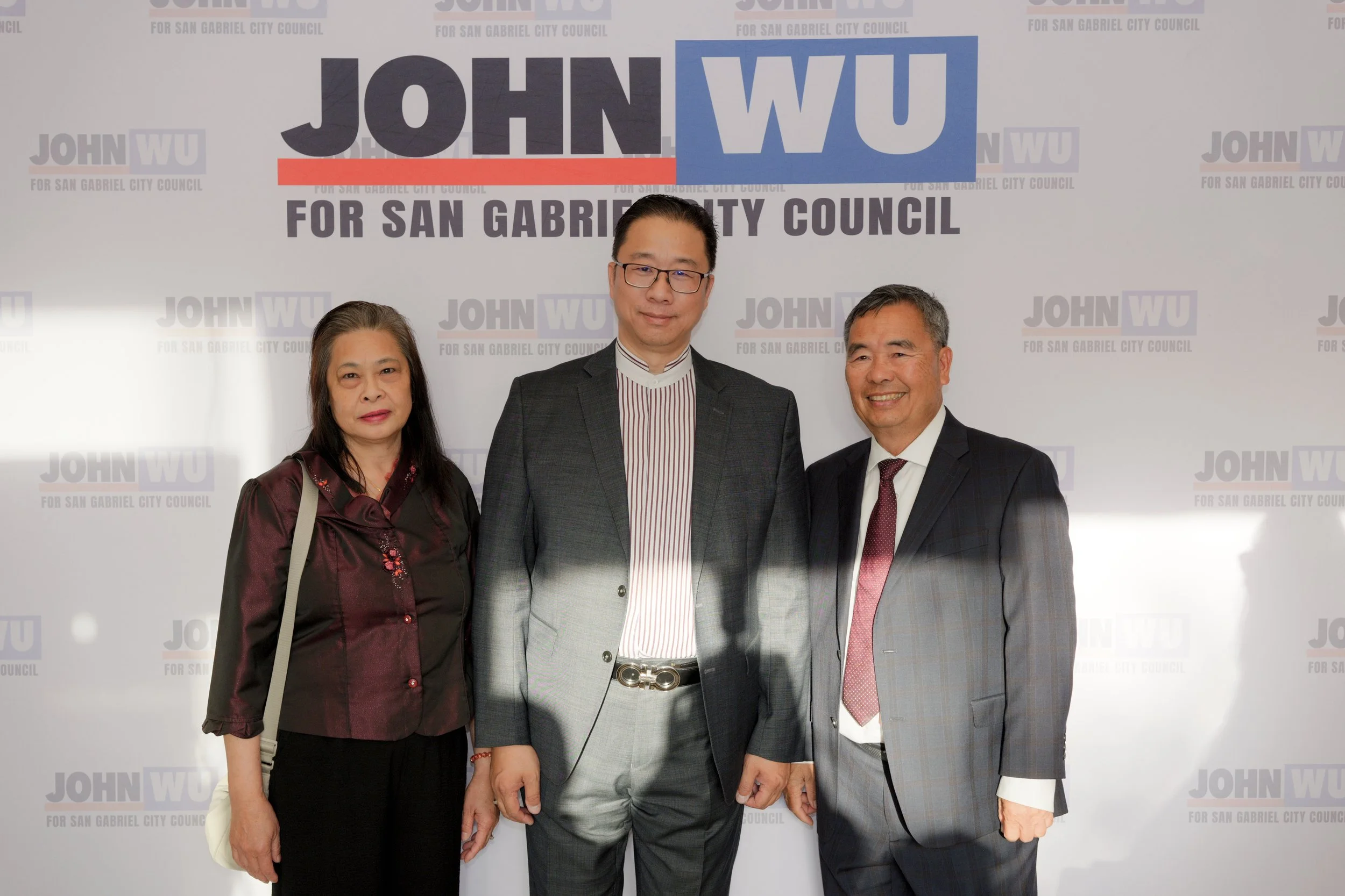 Three people standing in front of a campaign banner that reads 'JOHN WU FOR SAN GABRIEL CITY COUNCIL'. The woman on the left has long black hair, wears a dark red blouse, and has a beige handbag. The man in the middle has short dark hair, glasses, an