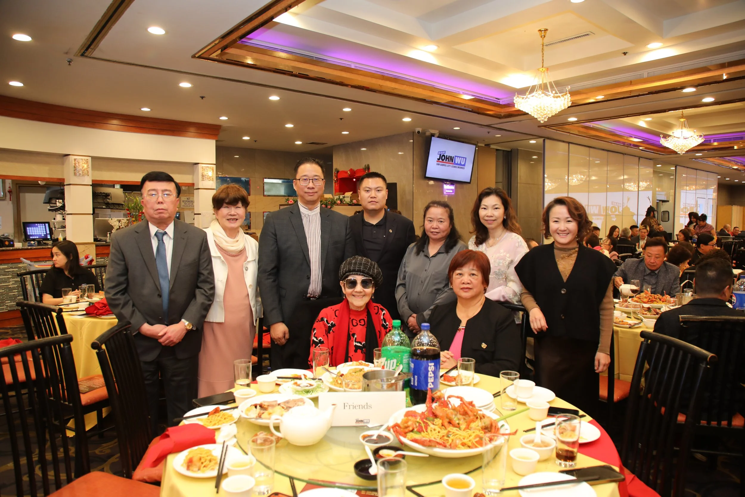 A group of people standing and sitting around a banquet table at a social gathering in a restaurant, with plates of food and drinks on the table and a large screen on the wall displaying a political campaign sign for John Wu.