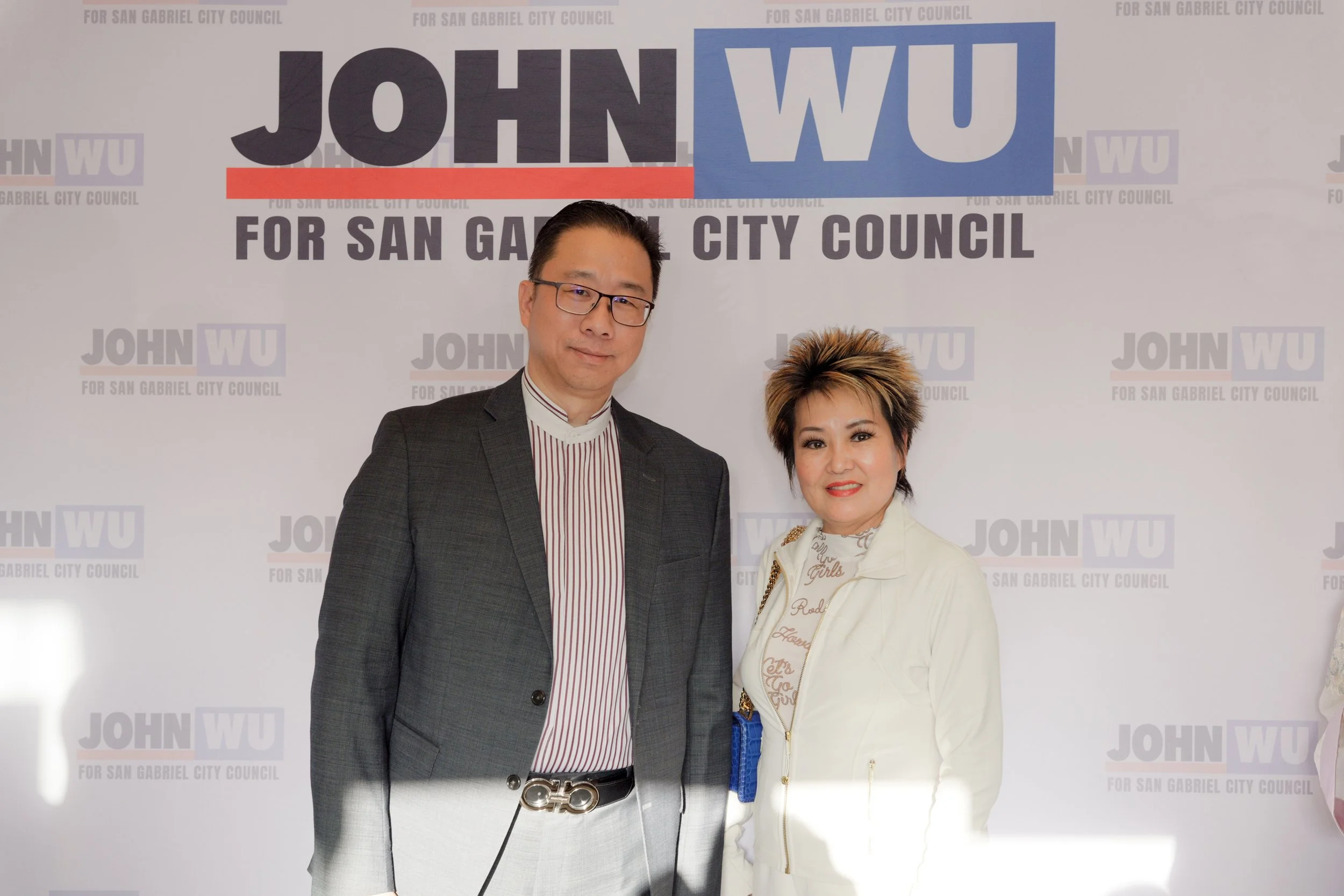 Two people, a man and a woman, standing in front of a step and repeat banner with the text 'John Wu for San Gabriel City Council' displayed repeatedly. The man is wearing glasses, a dark blazer, a striped shirt, and a belt with a prominent buckle. Th