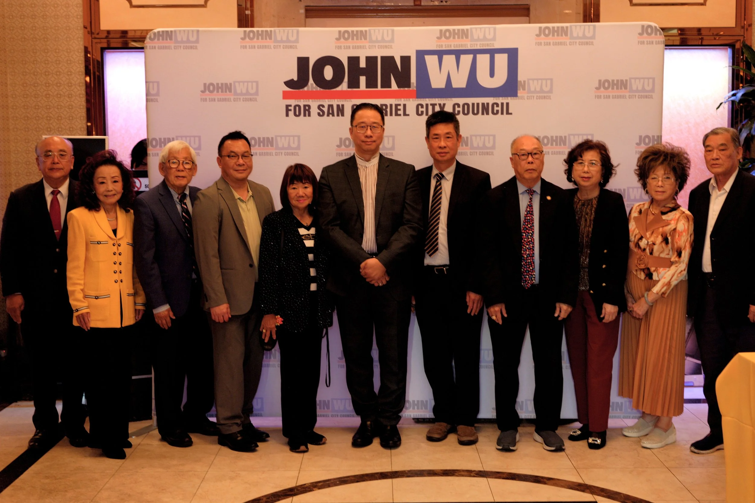 Group of diverse people standing in front of a backdrop with political campaign signs for John Wu for San Gabriel City Council.
