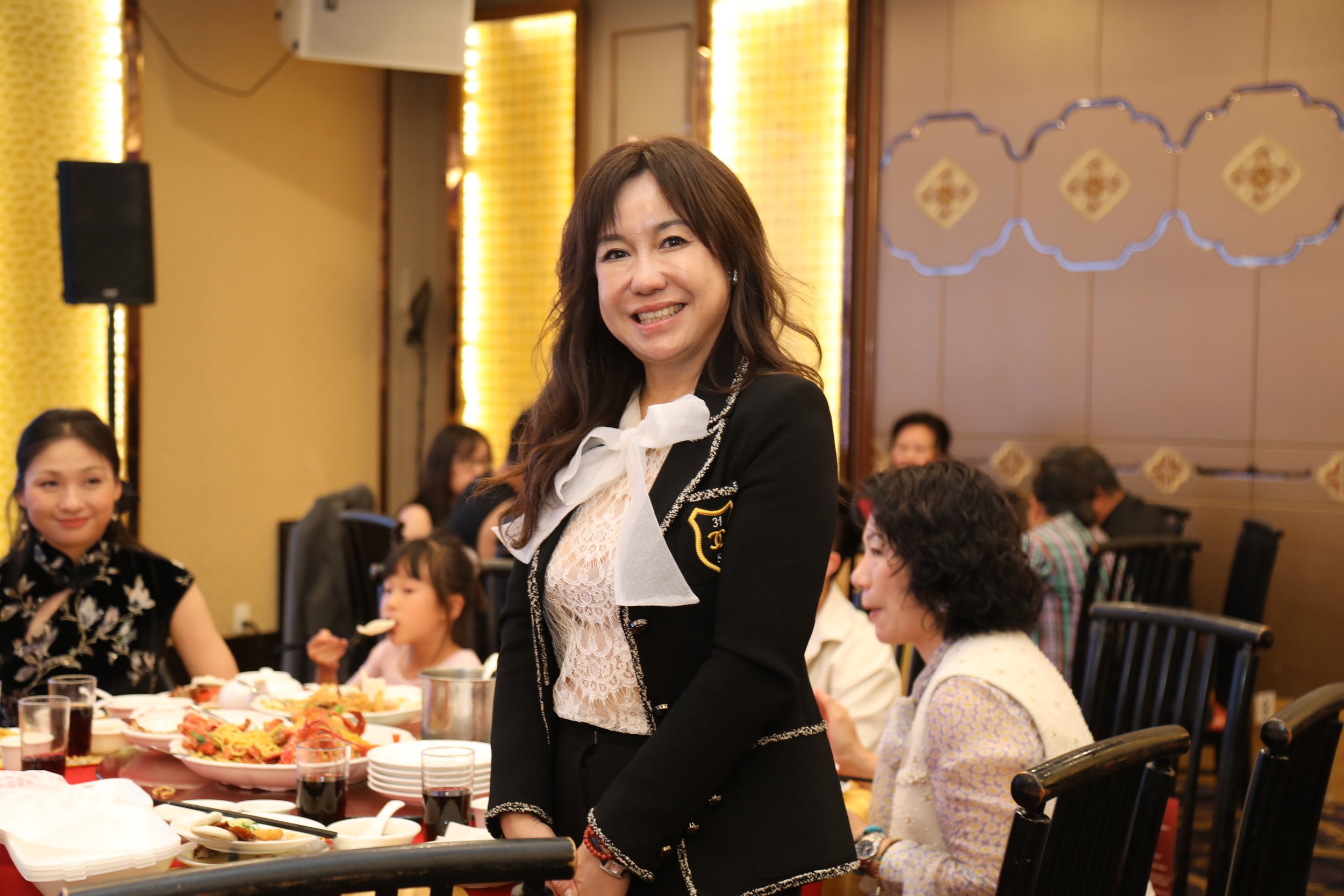A woman with long dark hair smiling at a social gathering seated at a table with food, surrounded by other people in a banquet hall with warm lighting and decorative wall panels.