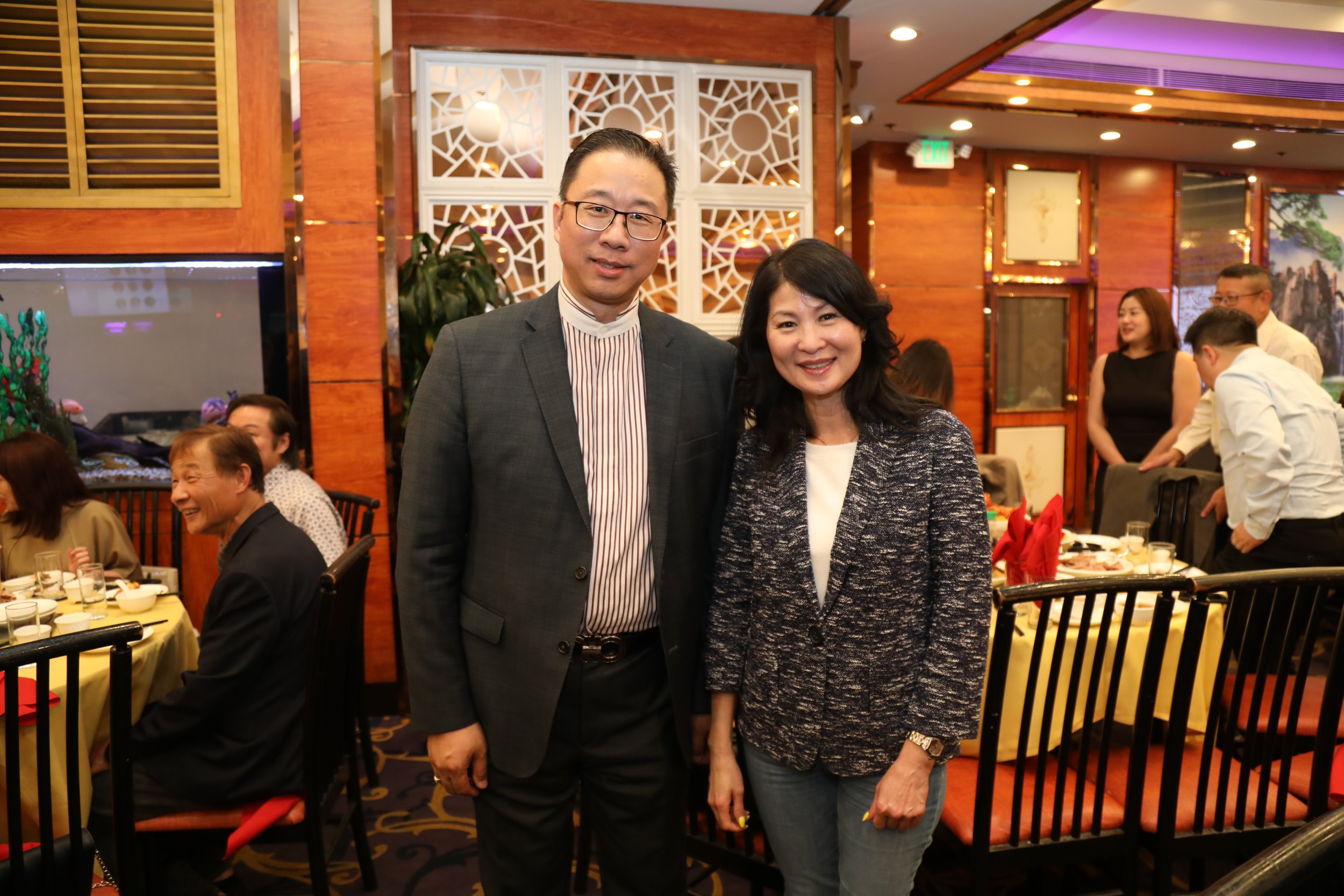 A man and woman standing together at a restaurant event, smiling and dressed in business casual attire, with other guests and a decorated dining area in the background.