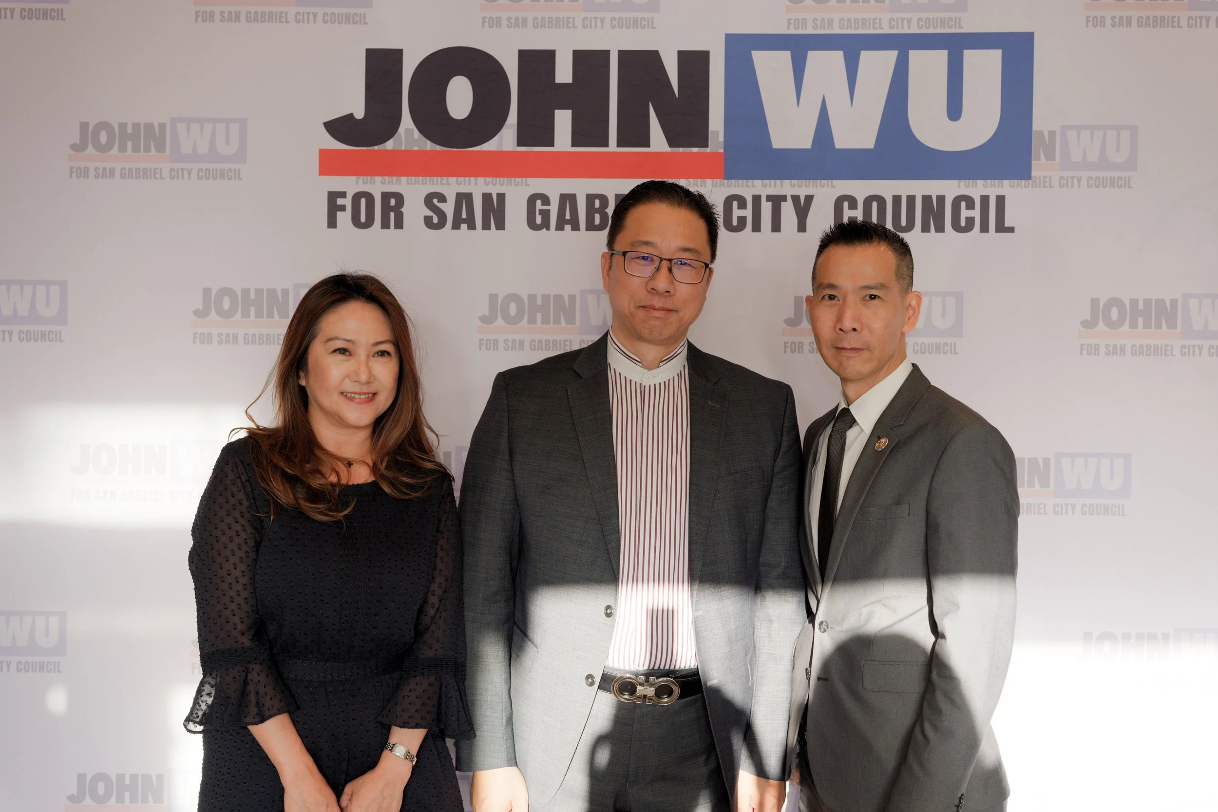 Three people standing in front of a backdrop with the text 'John Wu for San Gabriel City Council' repeated in the background. The woman on the left has long hair, is smiling, and wearing a black dress. The man in the middle has glasses, is dressed in