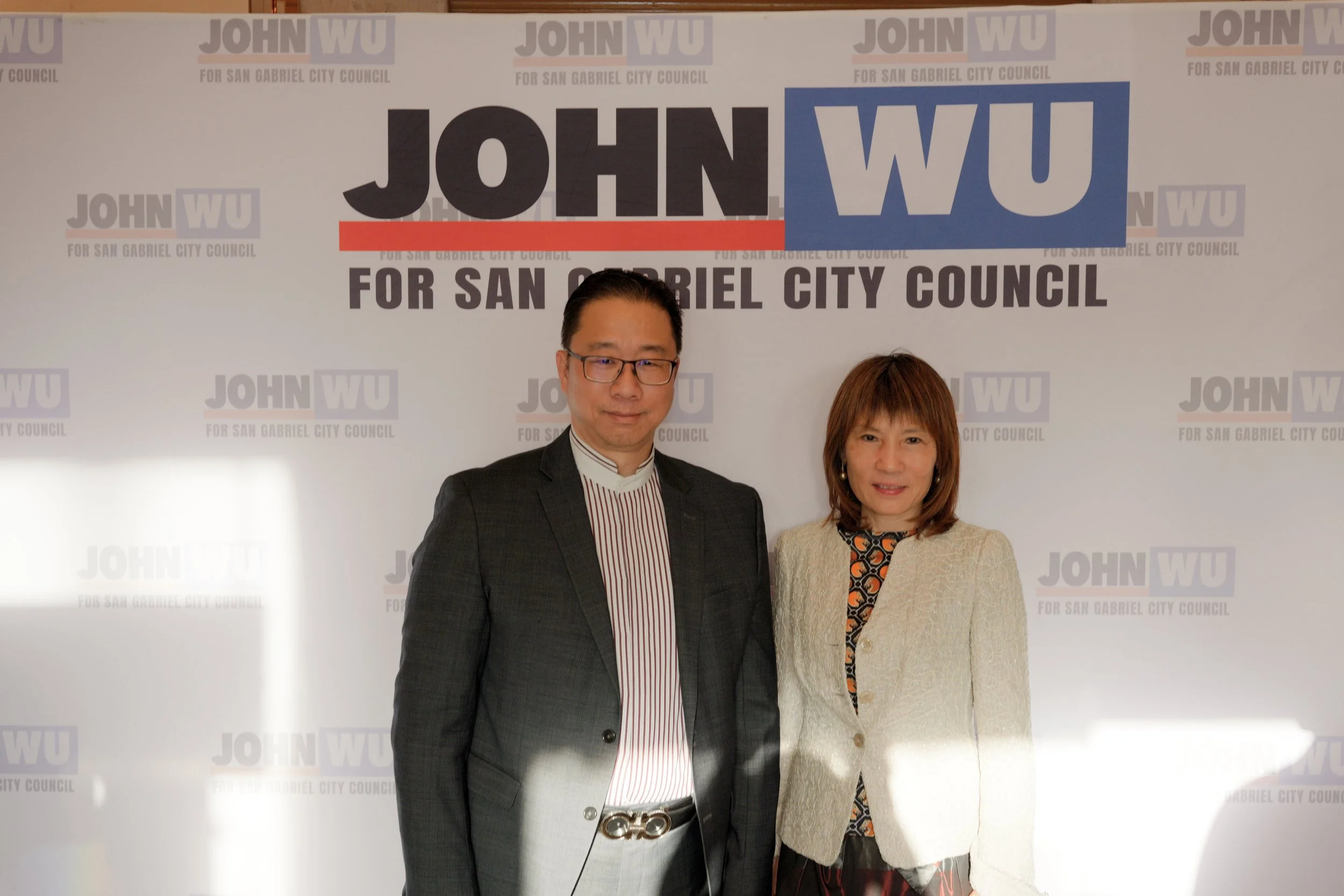 Two people, a man and a woman, standing in front of a campaign backdrop for John Wu for San Gabriel City Council. The man is wearing glasses, a dark blazer, and a striped shirt, while the woman has shoulder-length hair and is wearing a patterned top 