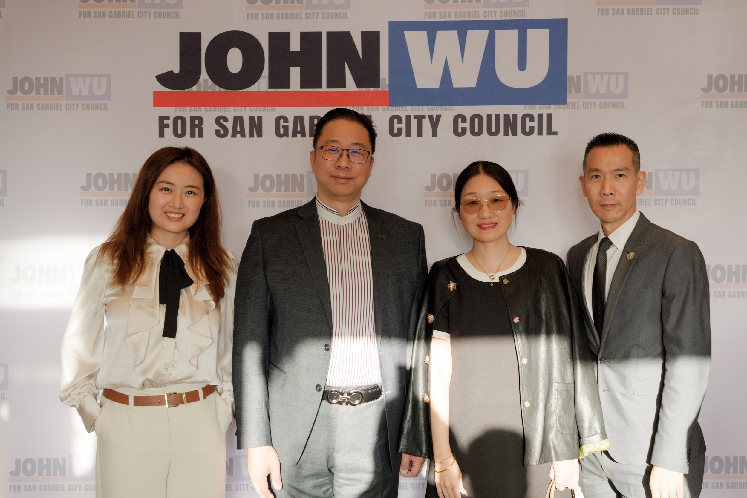 A group of five people are standing in front of a backdrop that has text supporting a candidate named John Wu for San Gabriel City Council. The group includes two women and three men, all wearing business attire, smiling and posing for the photo.