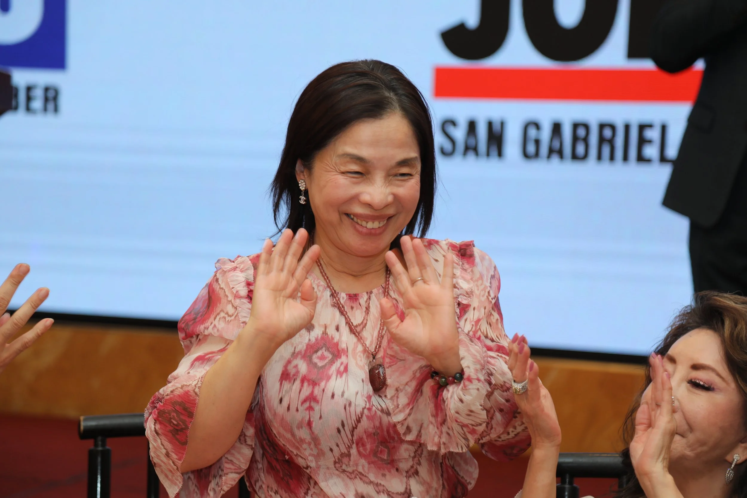 A woman with dark hair wearing a pink patterned top and jewelry, smiling and waving her hands. Behind her is a background with a logo and the text 'SAN GABRIEL'.