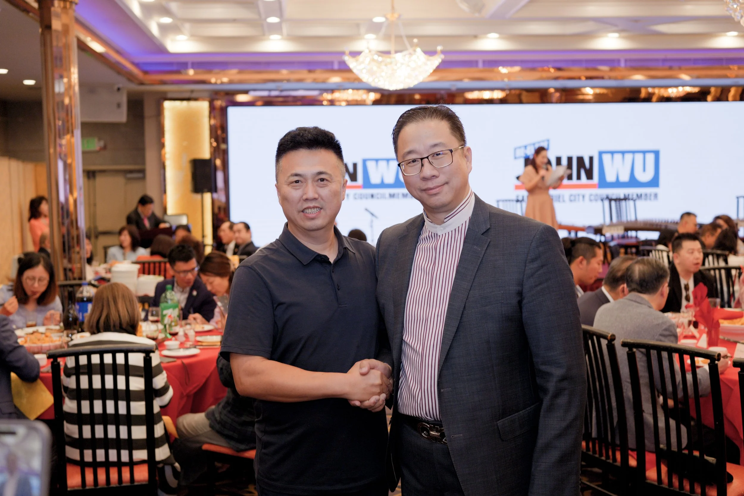 Two men shaking hands and smiling at a formal event, with a large screen in the background displaying the words "Western WU." The room is busy with multiple people seated at tables, eating and chatting, with a woman on stage giving a speech.