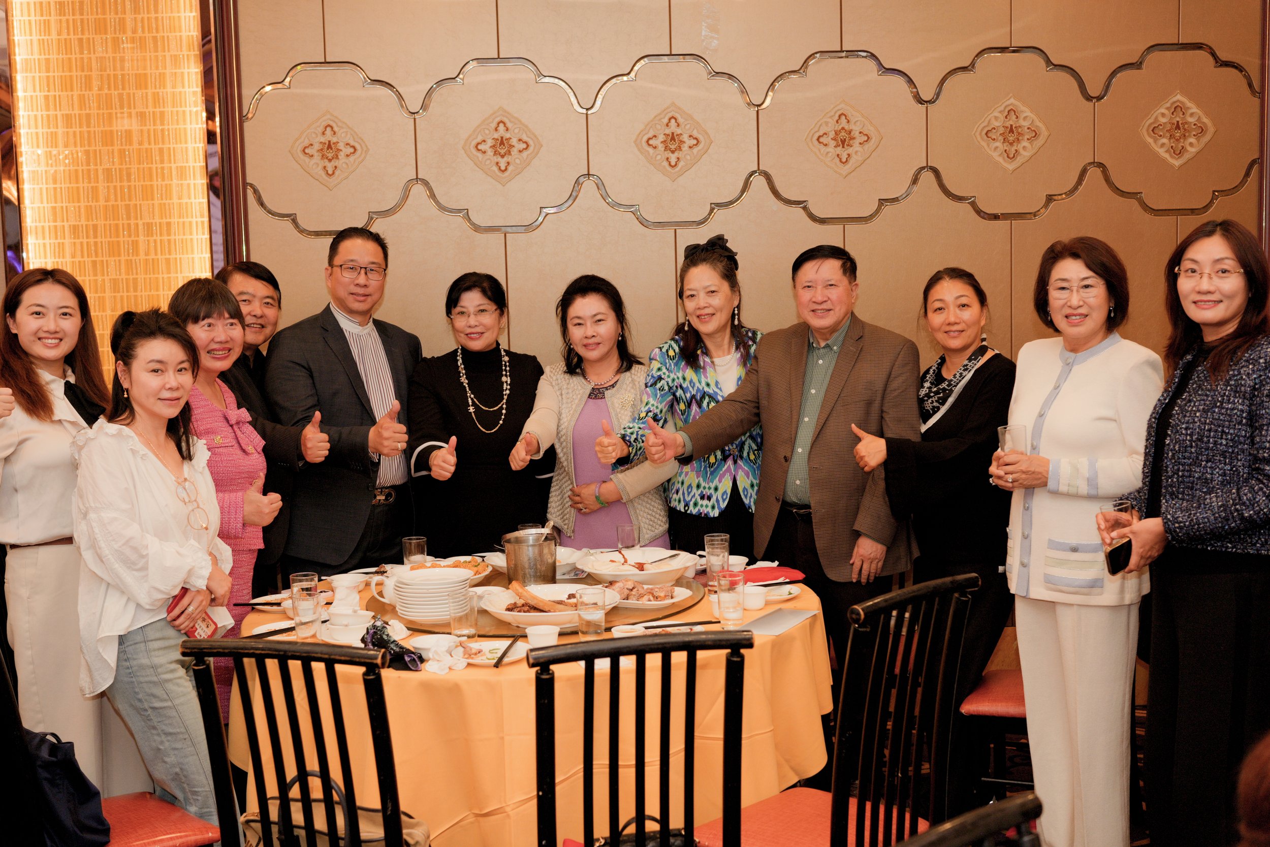 A group of people standing around a banquet table at a celebration, giving thumbs up, in a decorated room with beige walls and patterned wallpaper.